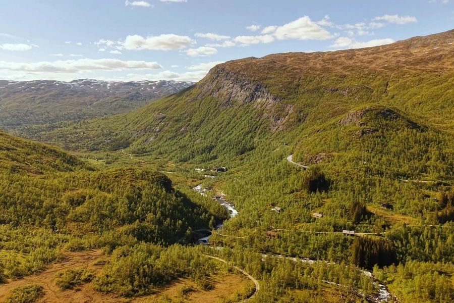 Mountain valley with a river, forested slopes, winding roads, and distant snow-dusted peaks.