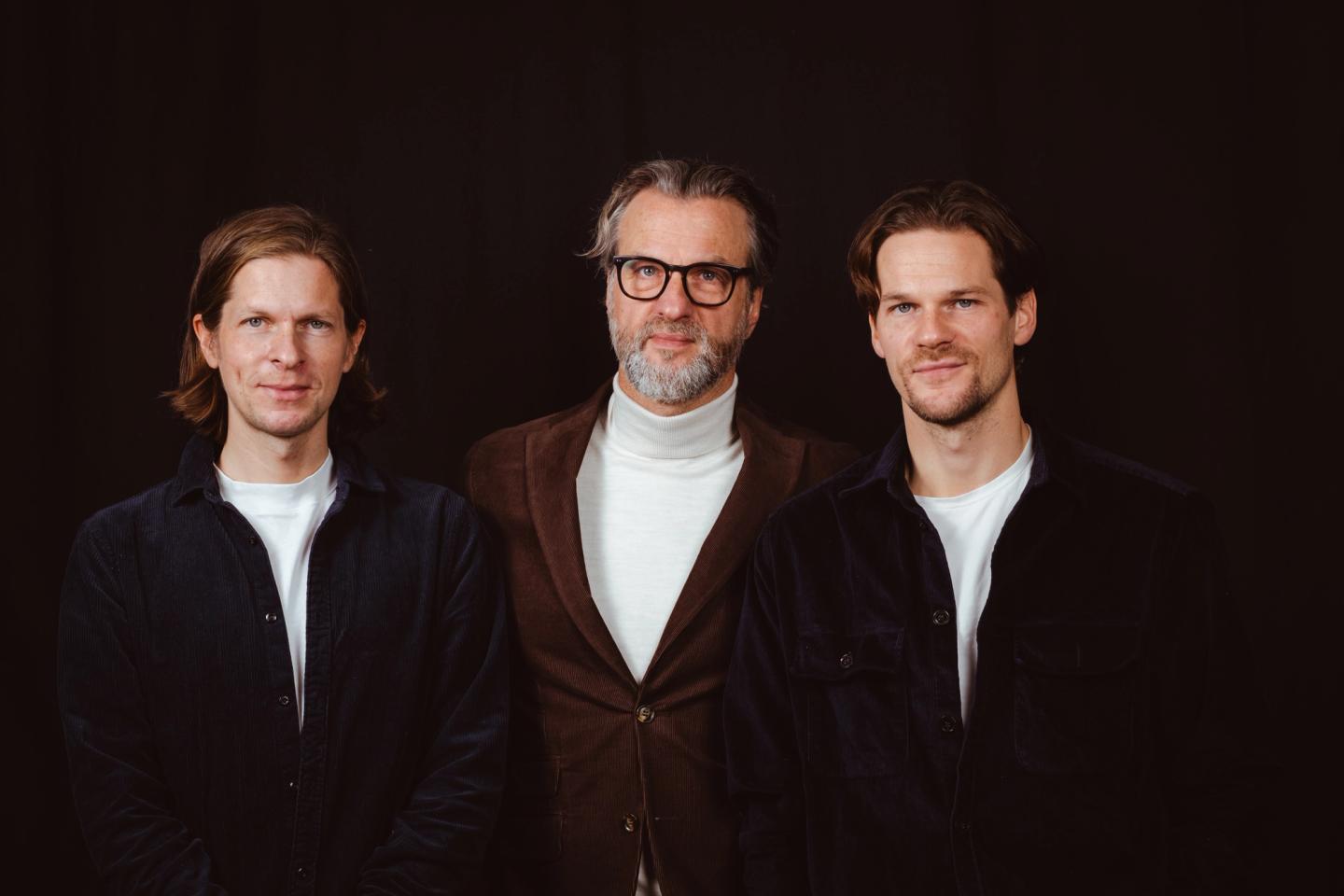 Tre menn som smiler. De to på hver side har mørkeblå skjorter mens han i midten har briller, grått skjegg og en brun blazer. 