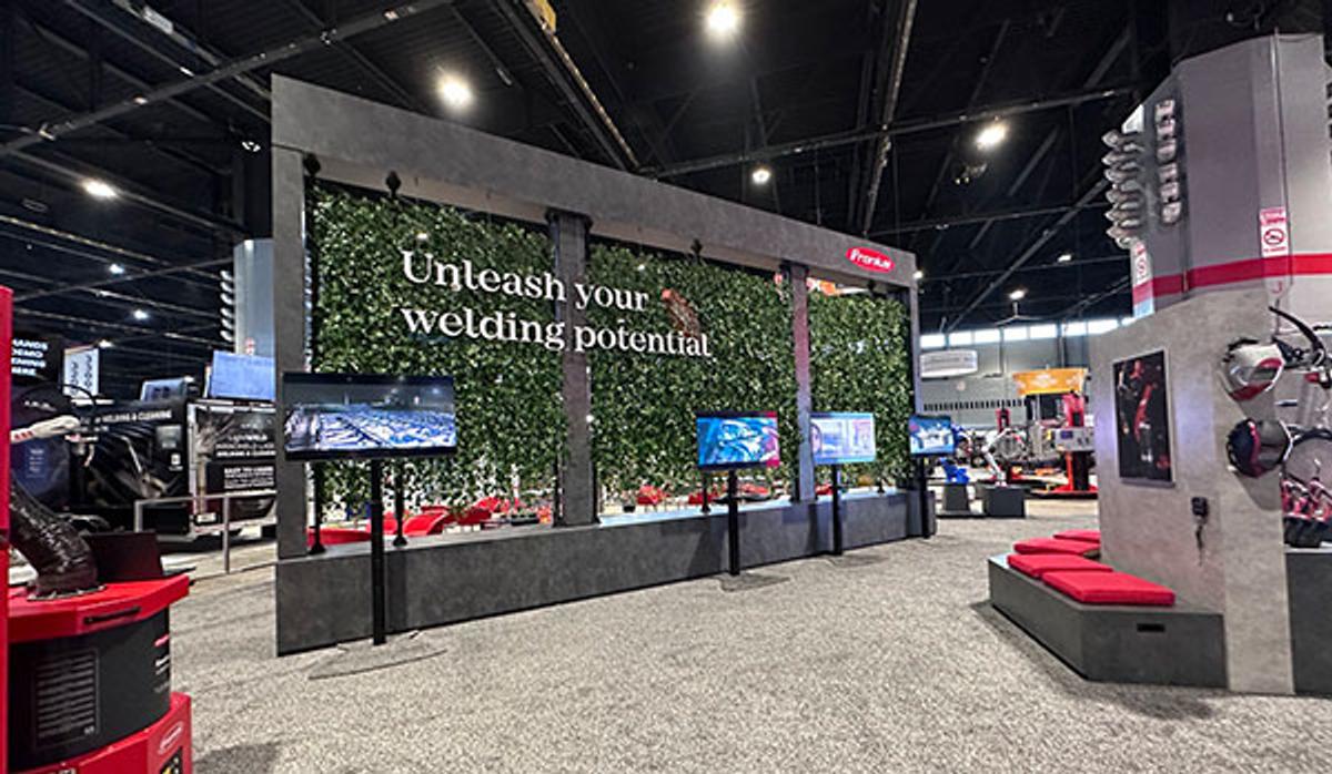 A tradeshow booth with a wall of green leaves, four TV screens, and words "Unleash your welding potential" 