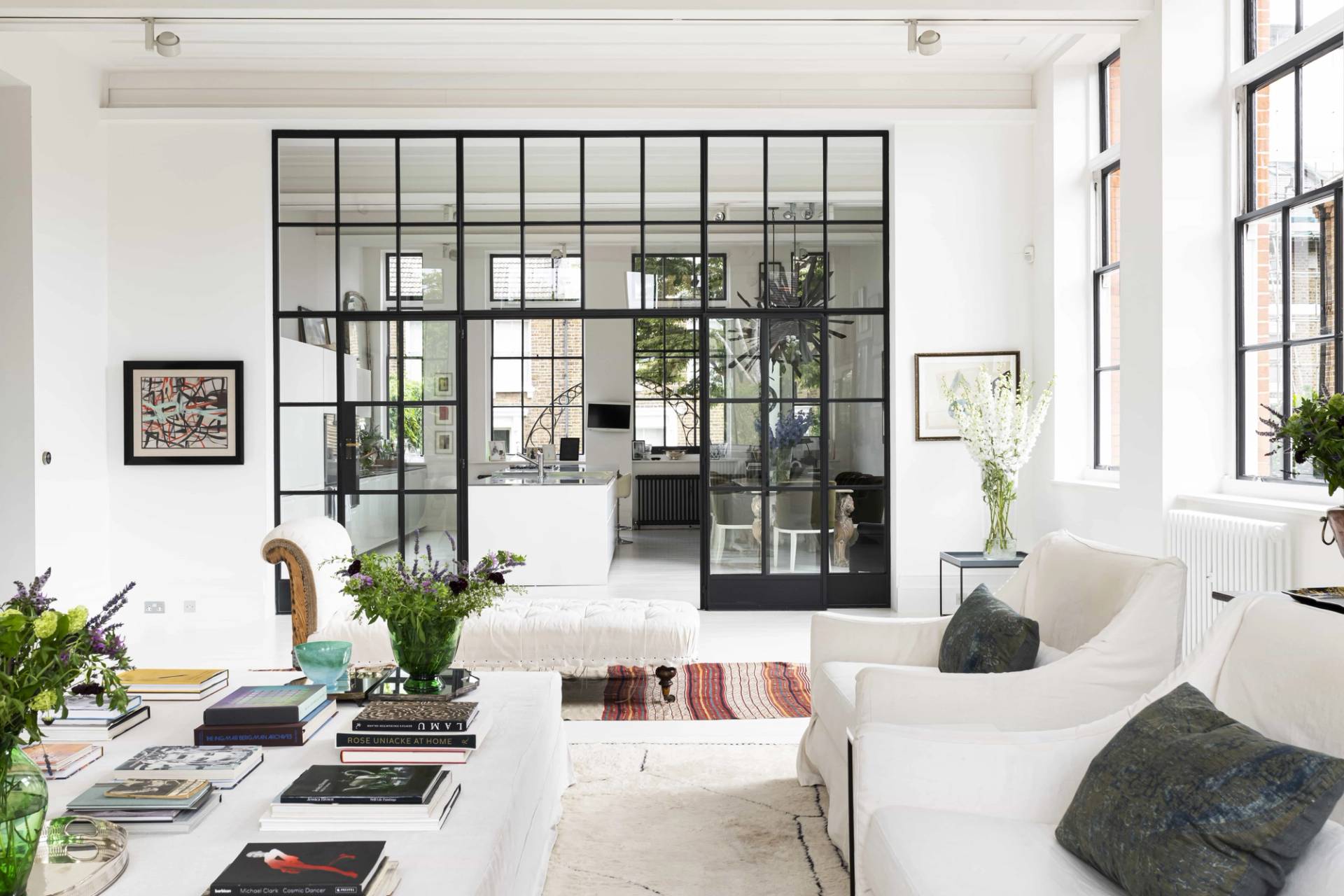 Symmetrical view through a black Crittall glass divider wall into a bright white living room with cream sofas and gallery wall art, Love Locations SW11.
