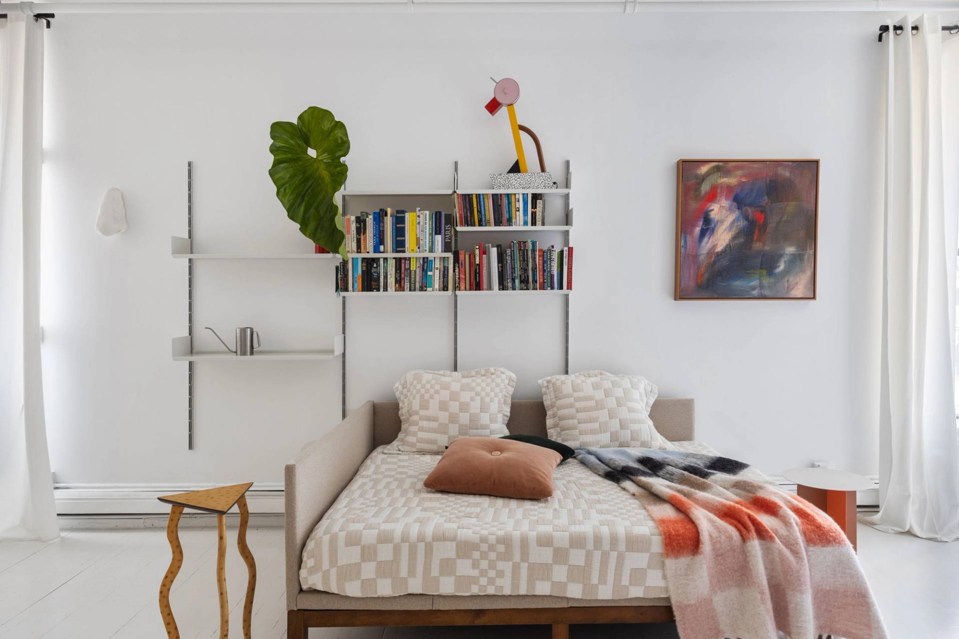 Artist bedroom photoshoot space with a minimalist bed, checkered orange blanket, wall-mounted bookshelf, and tropical potted plant in Tribeca 10007 from Love Locations.