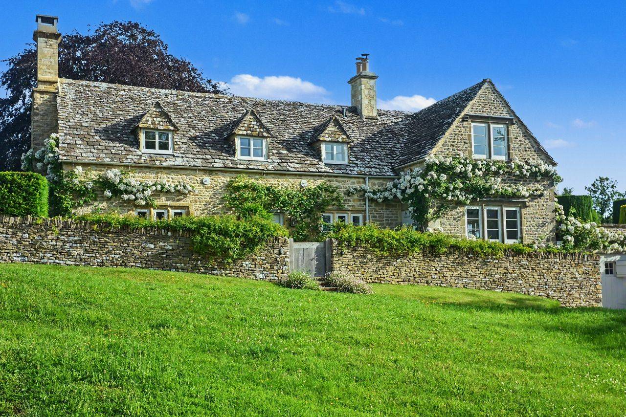 Historic stone cottage with climbing greenery photoshoot location in Gloucestershire GL7 through Love Locations, featuring a low drystone wall and sprawling lawn.
