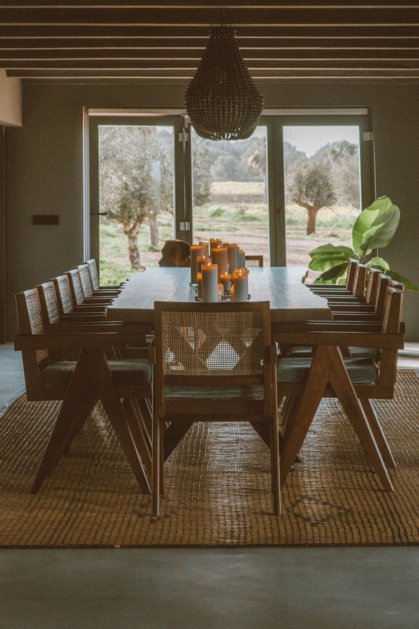 Sunlit rustic dining space set with wooden chairs, green indoor plants, and view of olive trees, Love Locations 07630.