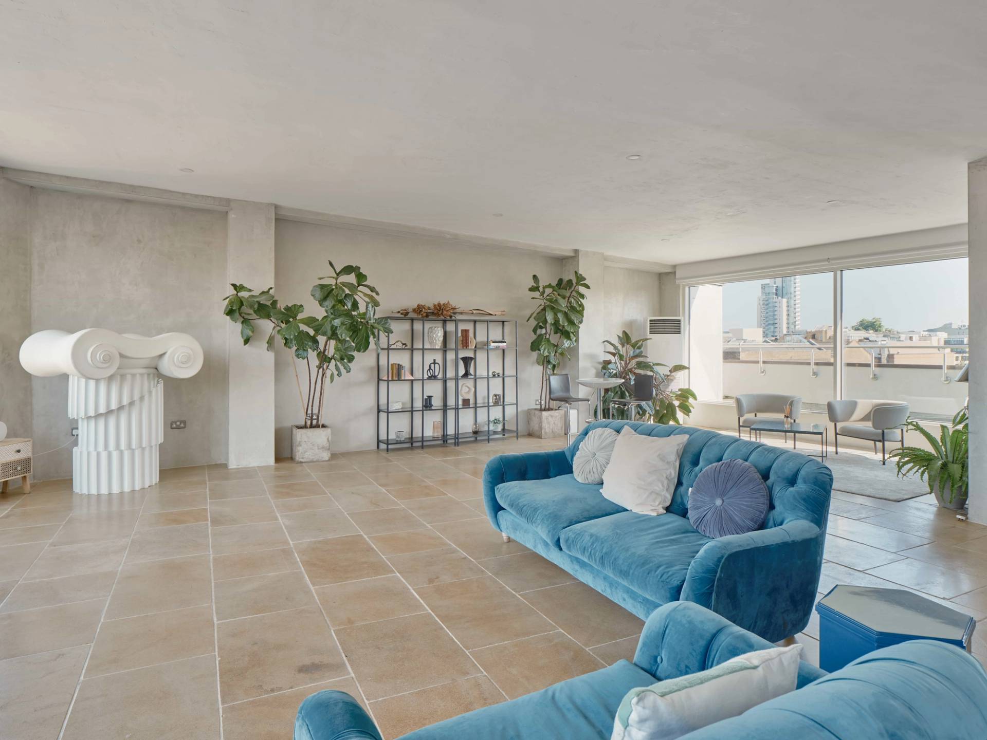Stylish open-plan penthouse lounge at THE PENTHOUSE SE1 featuring blue velvet sofas, modern decor, large windows with city views, and contemporary art and plants.