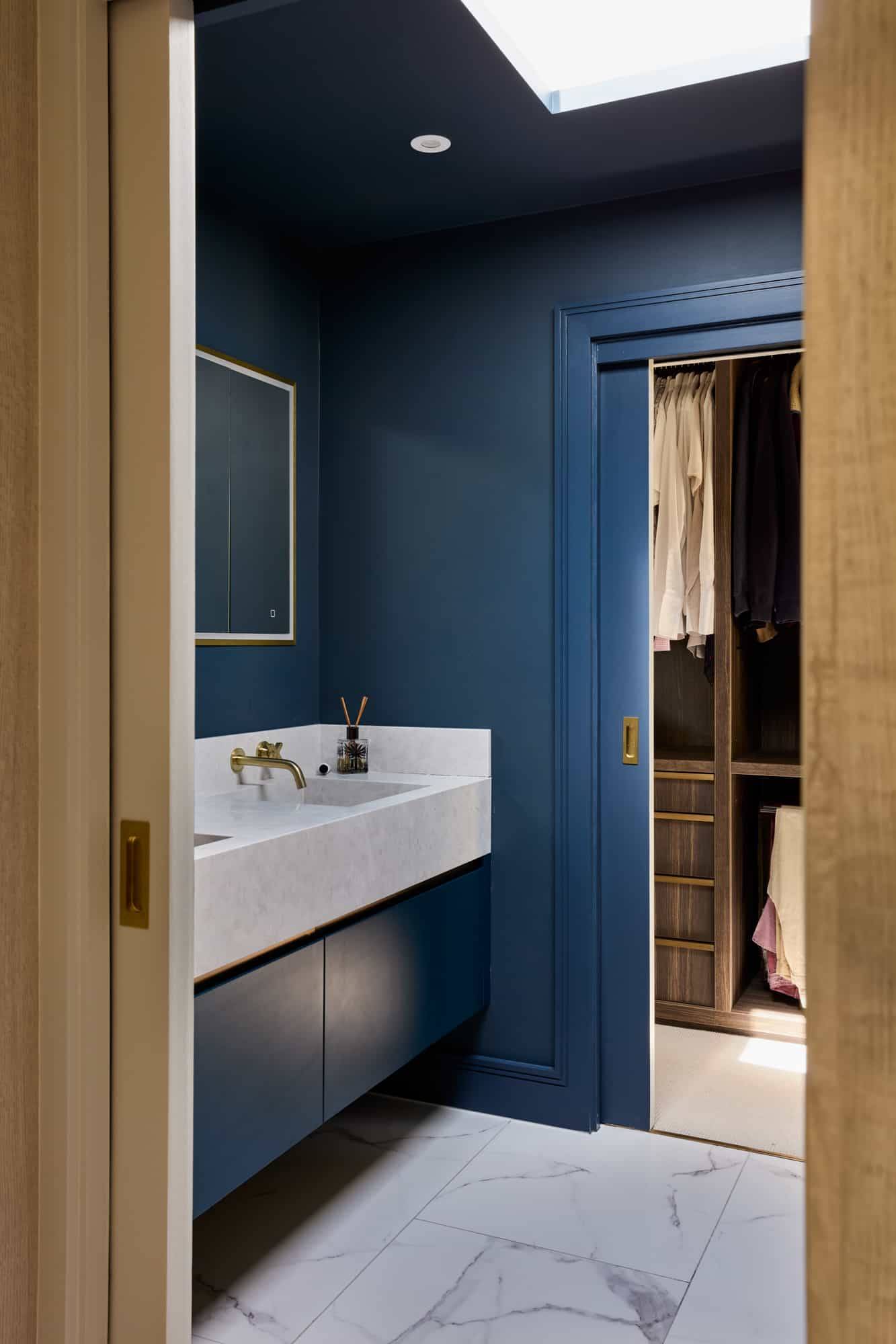 Deep blue ensuite bathroom featuring a white double sink vanity, gold taps, and access to a dressing closet, Love Locations.
