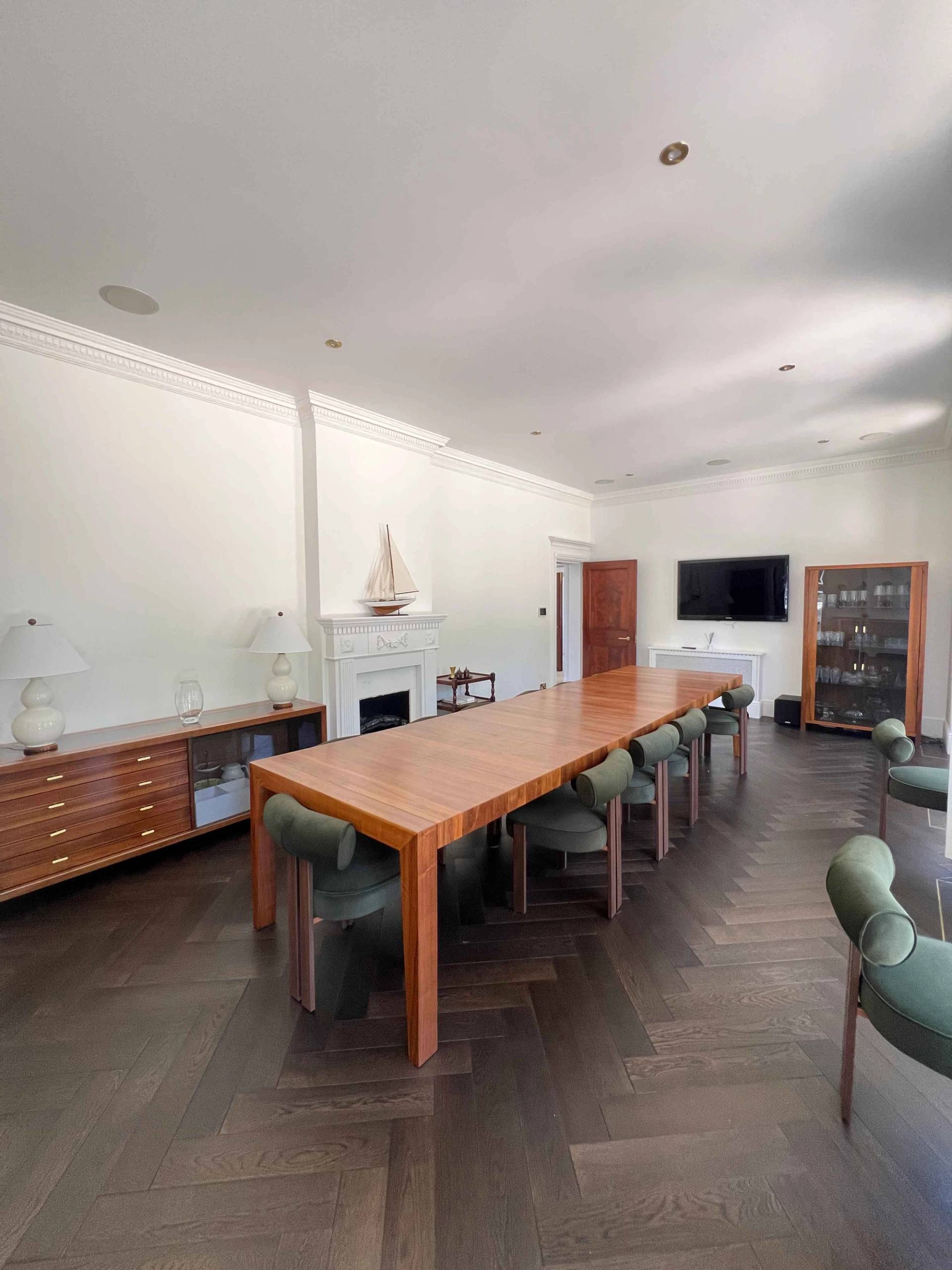 Wide-angle perspective of a long wooden dining table set on dark parquet floorboards under ambient ceiling lights, Love Locations BR7.