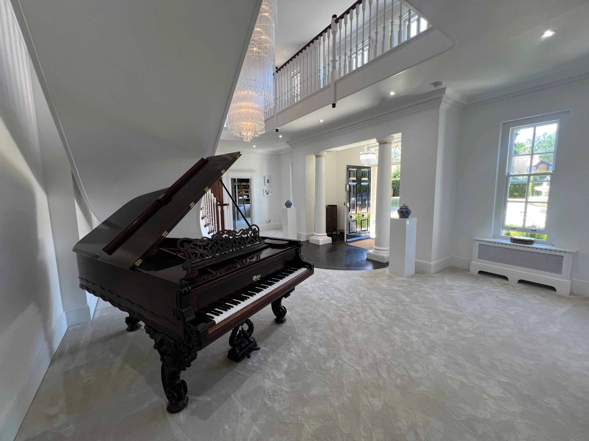 Black grand piano positioned in a double-height entrance hall beside a curved white staircase, Love Locations BR7.