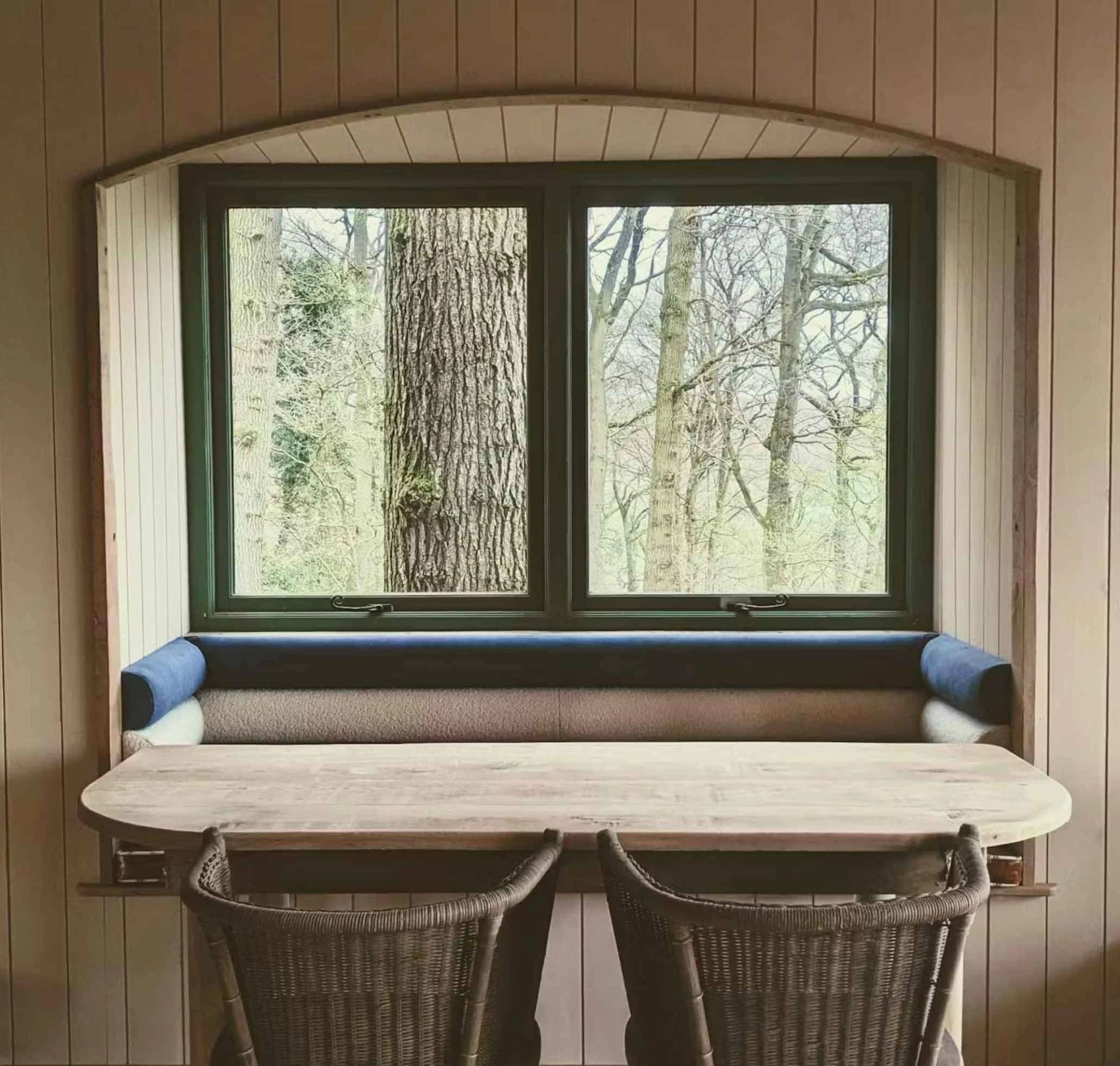 Bespoke wooden dining table and bench seating arranged perfectly in front of a large window viewing a massive tree trunk, unique production set via Love Locations.