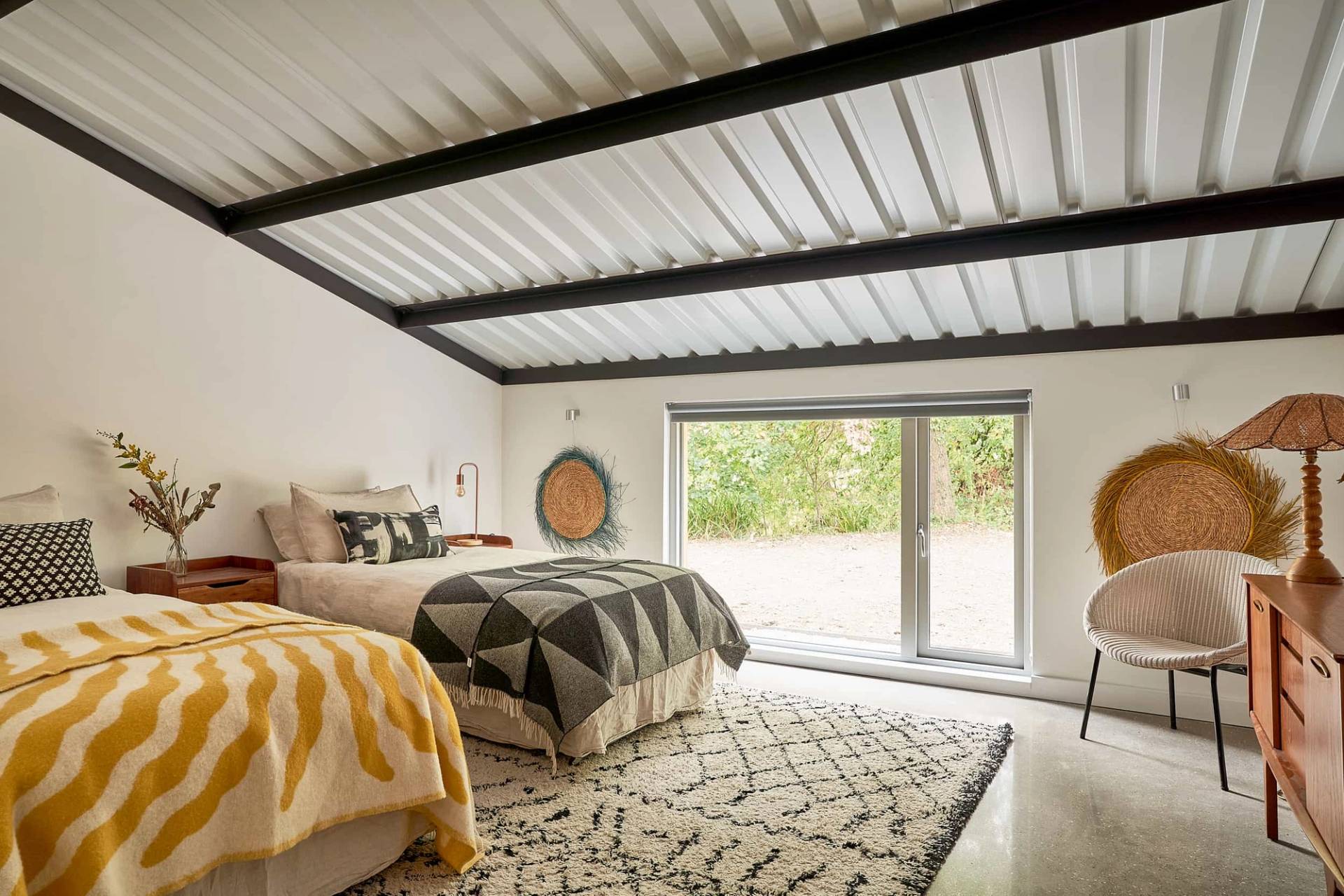 Guest bedroom featuring twin beds with mustard yellow throws, industrial metal roof ceiling, and glass sliding door, Love Locations IP30.