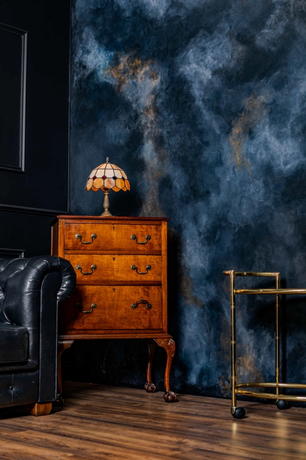 Vintage wooden chest of drawers set against a dramatic dark blue and gold clouds backdrop with a Tiffany-style lamp, Love Locations N15.