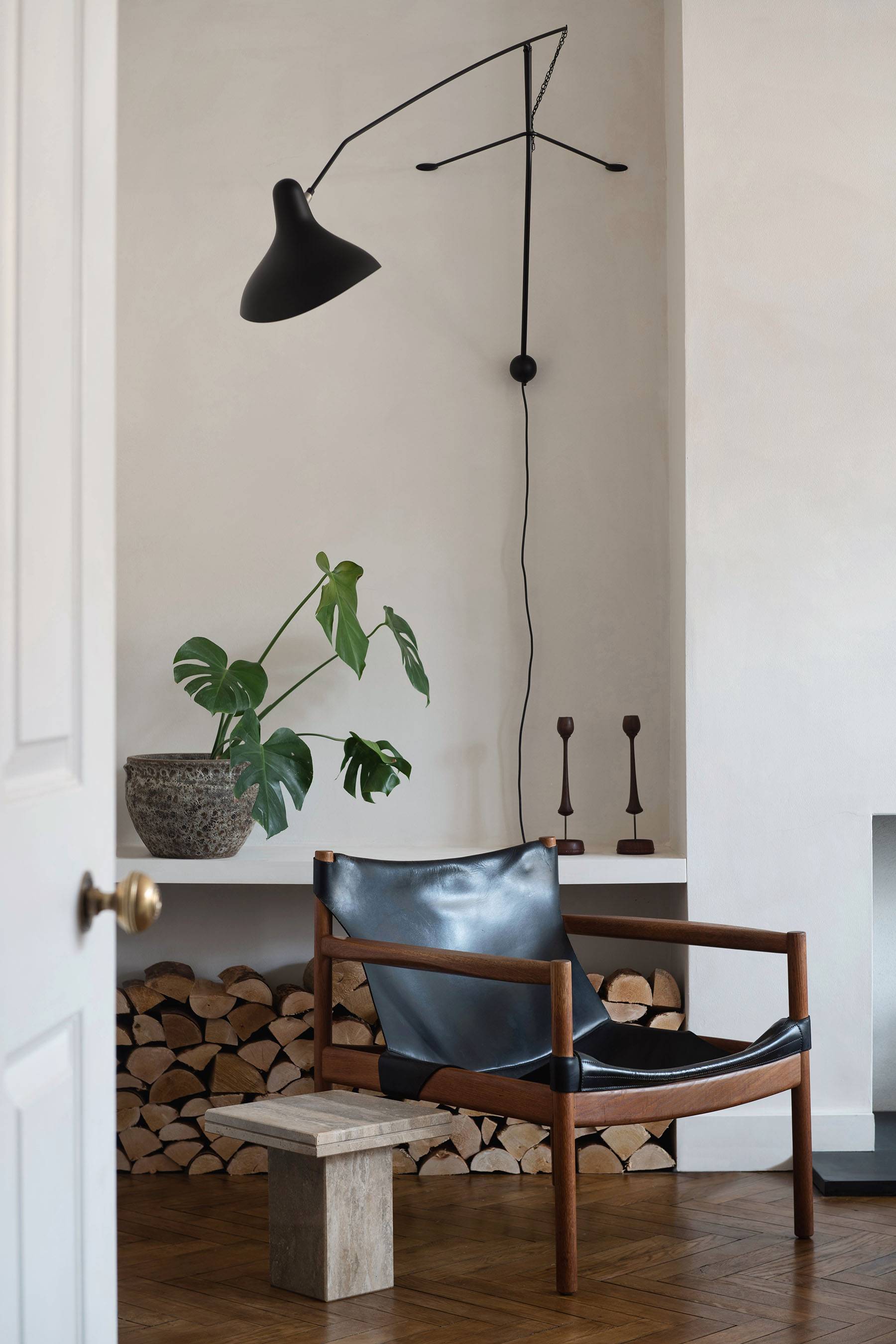 Minimalist plaster wall fireplace alcove with stacked firewood, dark armchair, and glass coffee table, Love Locations SW16.
