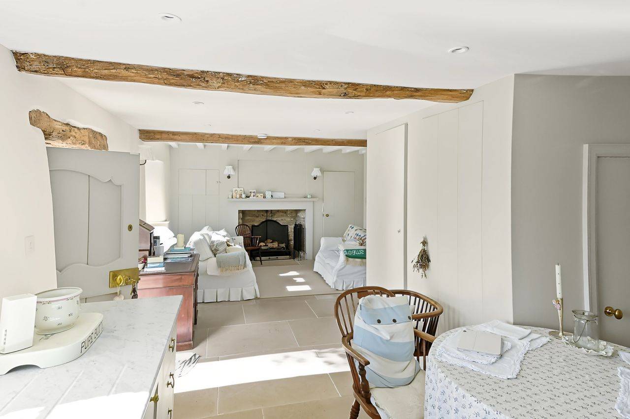 Bright cottage living room film location in Gloucestershire GL7 from Love Locations, featuring white walls, exposed timber ceiling beams, neutral sofas, and an inglenook fireplace.