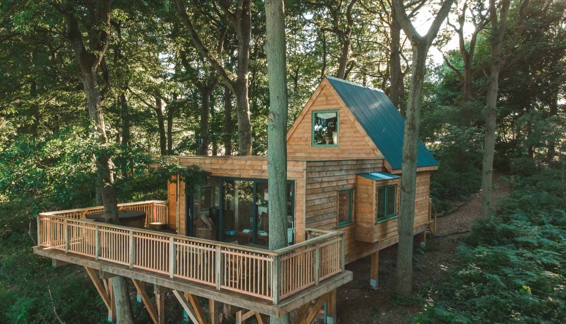 High-angle exterior look at a modern architectural timber treehouse built directly into a dense green woodland canopy, managed by Love Locations.