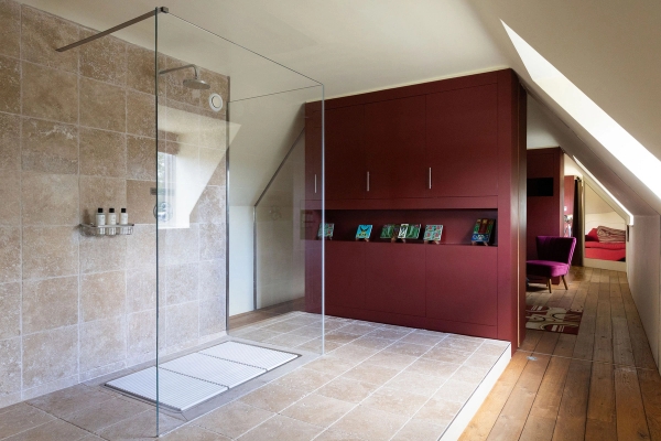 Love Locations luxury travertine bathroom West Sussex BN44: Architectural walk-in shower room lined with beige stone tiles, glass partition screen, and a bold deep-red storage partition wall.