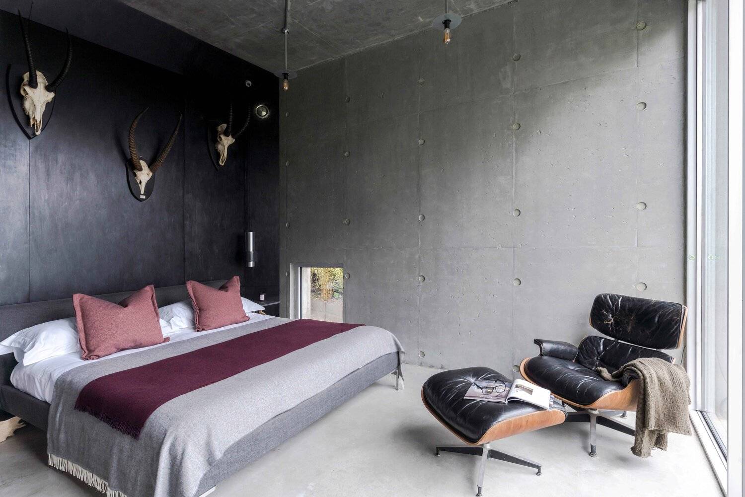 Master bedroom suite set against brutalist concrete walls featuring an Eames lounge chair, ottoman, and dark feature wall, Love Locations BN7.