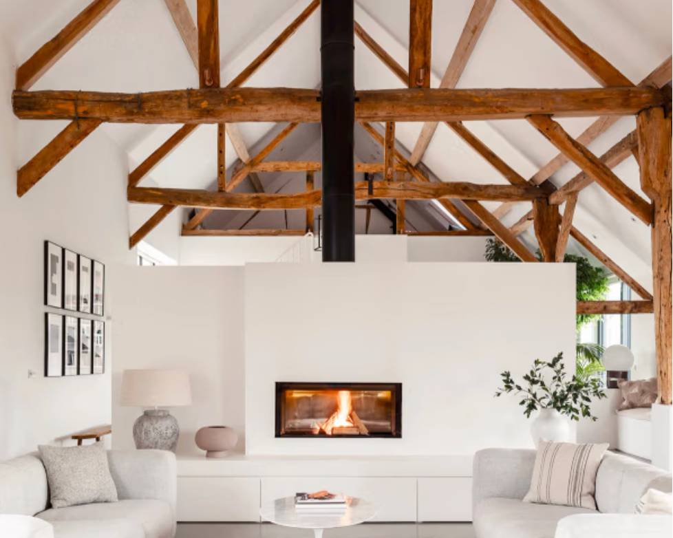 Open-plan dining and living space at Ham House with exposed timber beams, a large wooden dining table, contemporary fireplace, polished concrete floor, and floor-to-ceiling glazing flooding the room with natural light.
