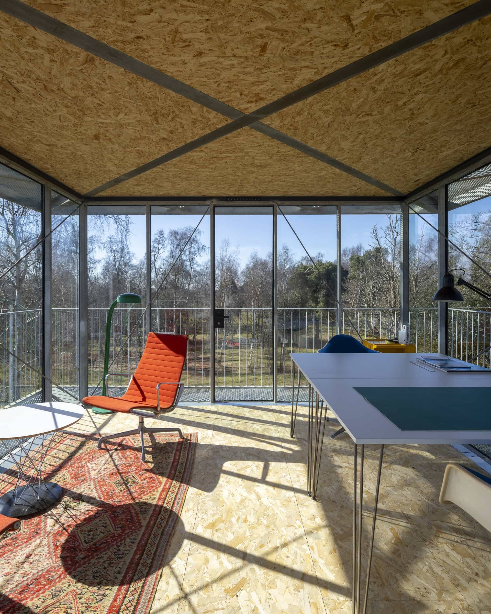 Sunlit elevated glass observation studio with red lounge chairs, rug, and desk area overlooking fields, Love Locations IP17.