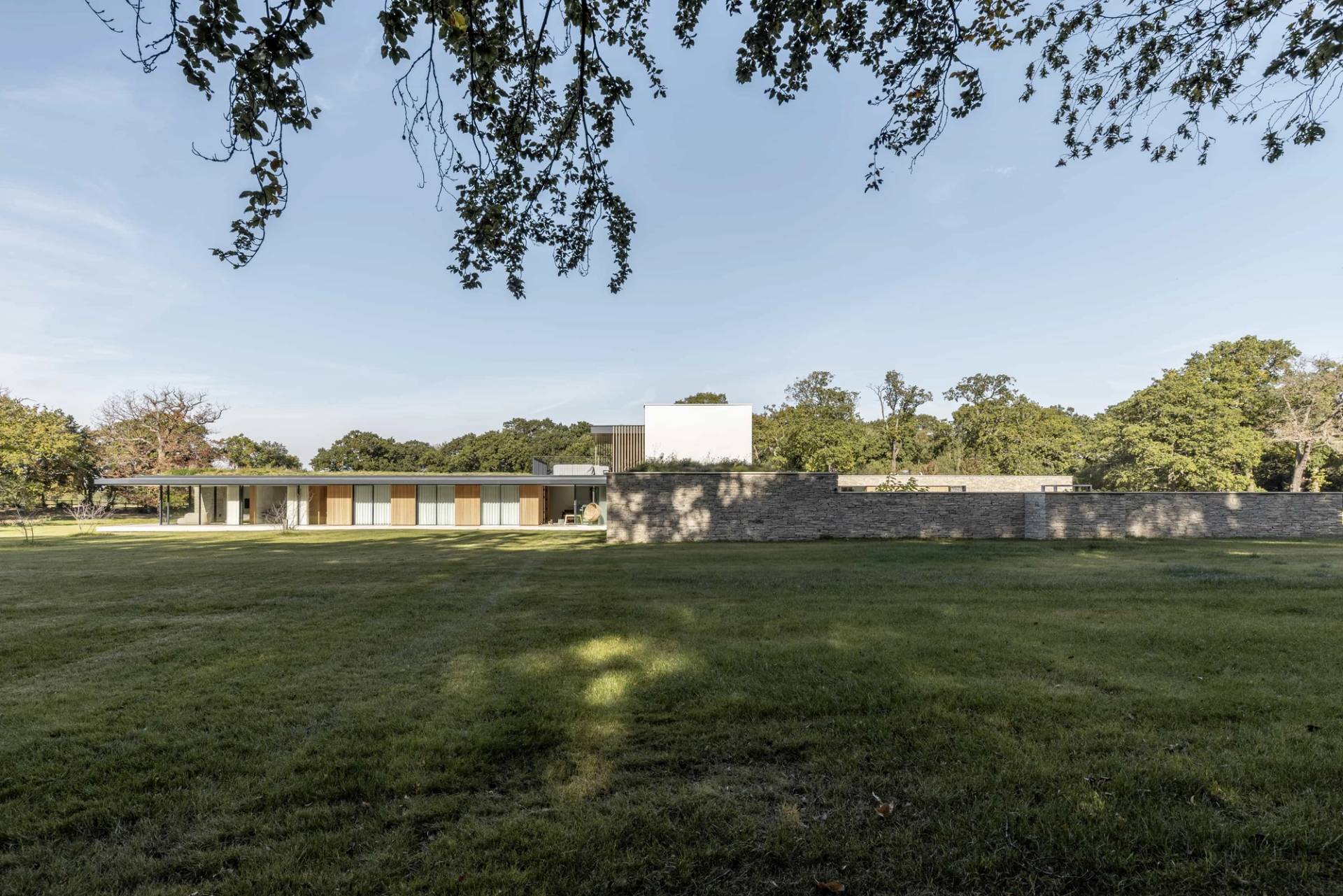 Distant rear view of a luxury minimalist new build property highlighting its horizontal profile, stone masonry, and vast open lawns, Love Locations.