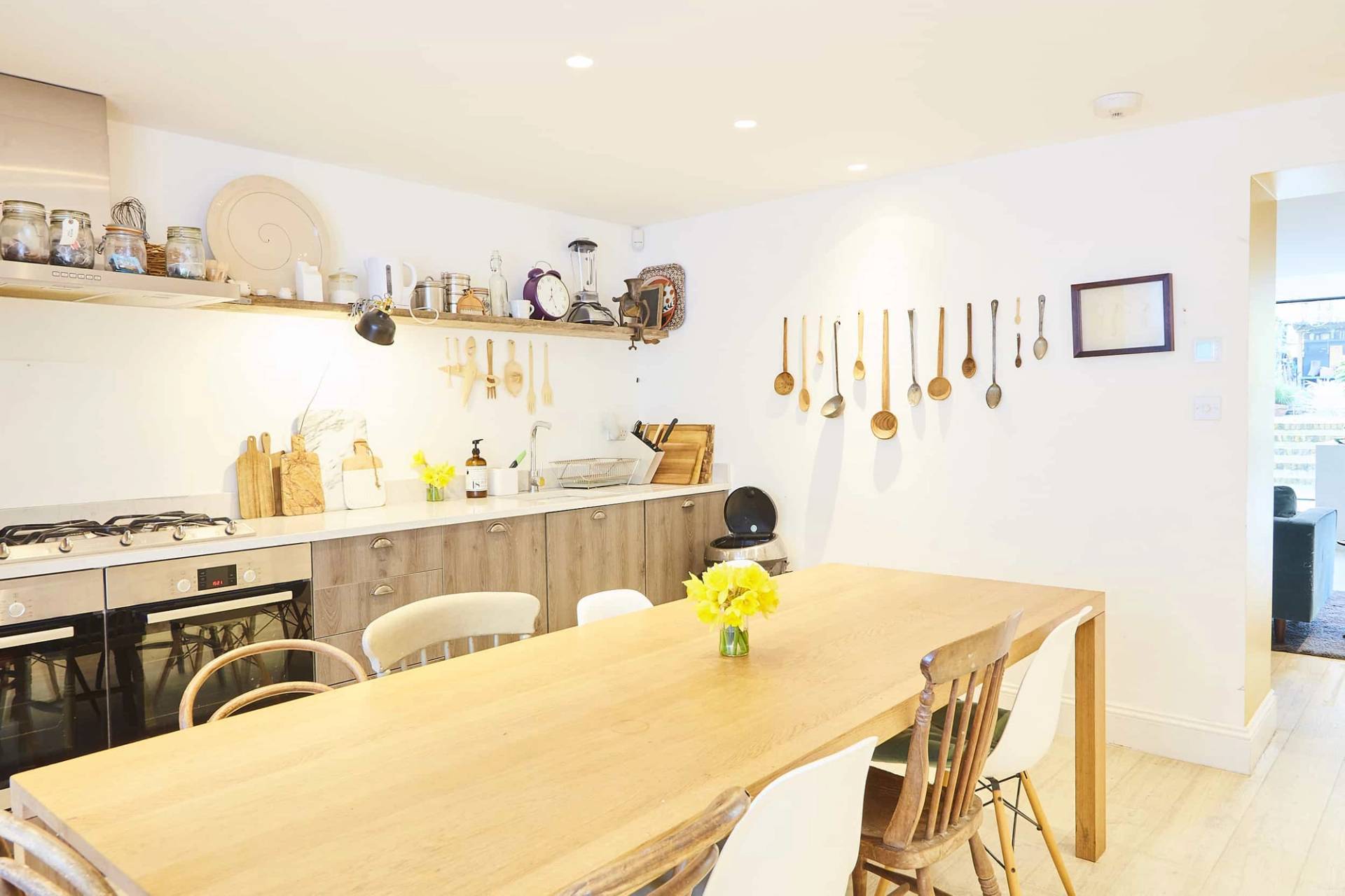 Warm dining room setup featuring a solid wood dining table, white dining chairs, and hanging wooden utensil wall display, Love Locations N5.