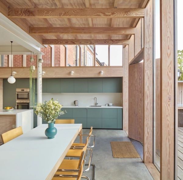 Open-plan dining space set beneath an exposed timber joist ceiling with green kitchen cabinetry, Love Locations E17.