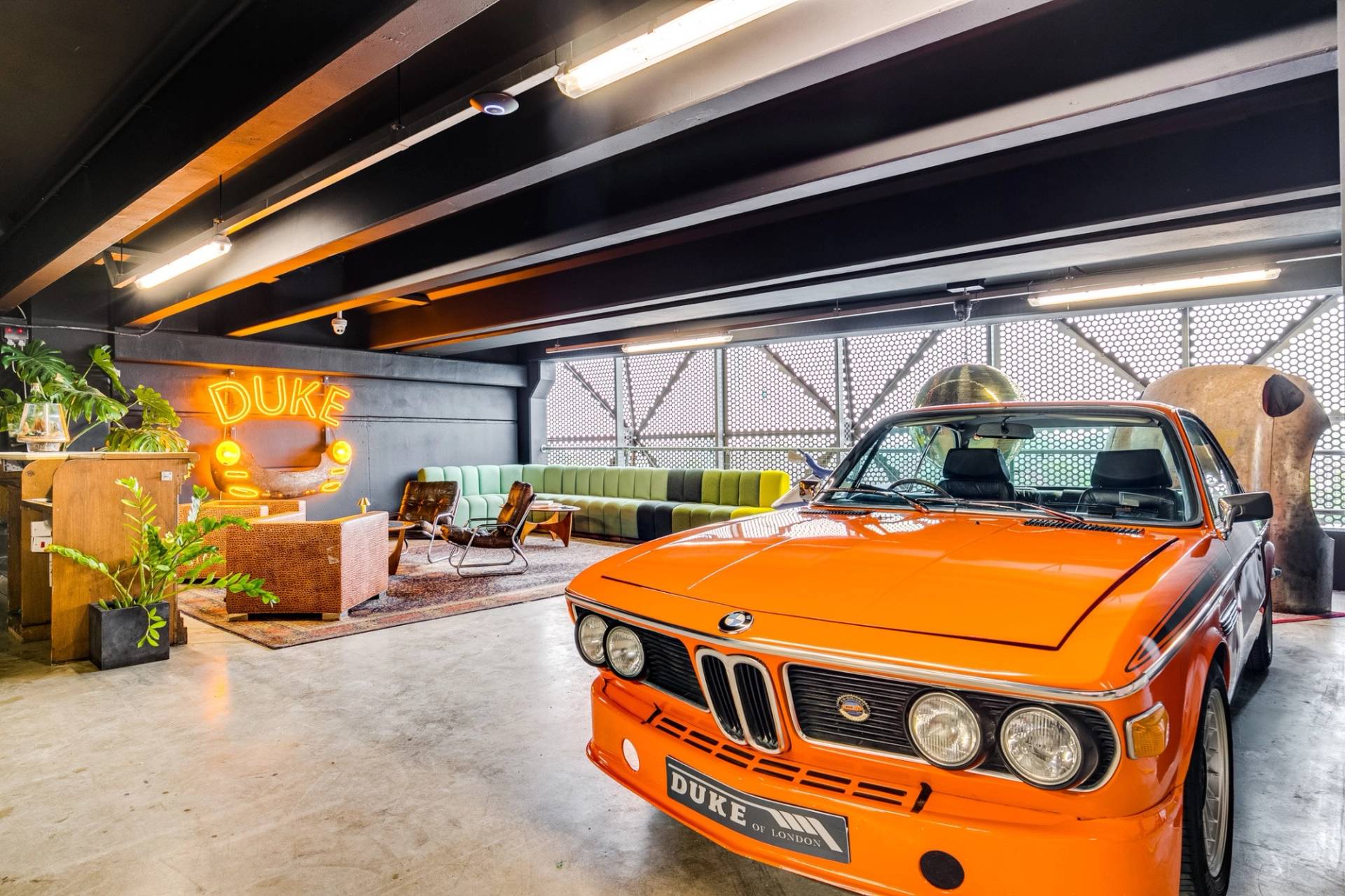 Classic orange BMW parked inside a retro 1950s car showroom film location in Brentford TW8 from Love Locations, featuring black industrial ceiling beams and warm DUKE neon lighting.