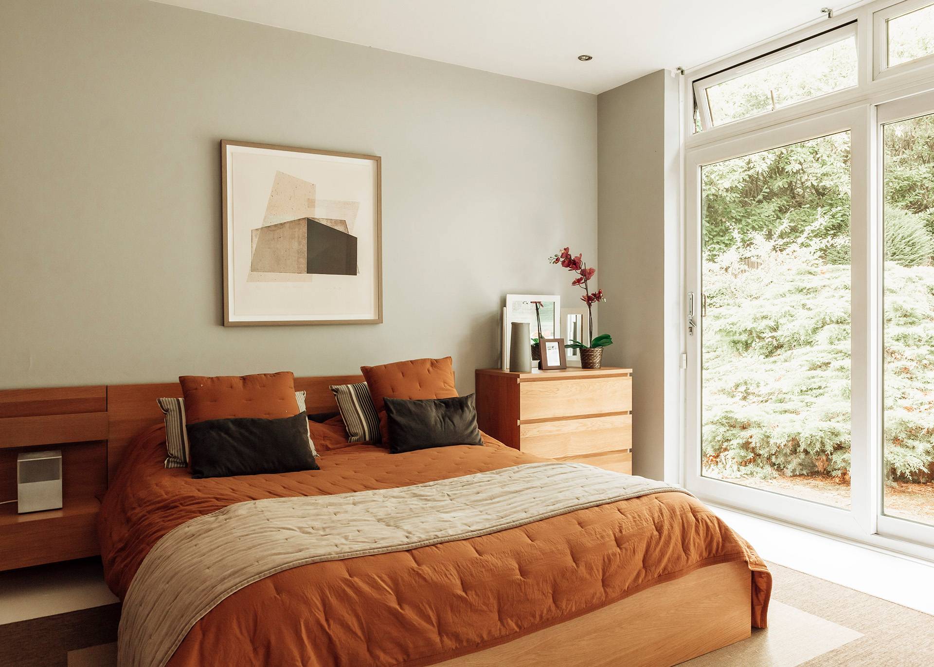 Bright modern guest bedroom featuring terracotta-toned bedding, warm wooden furniture, and tall windows, Love Locations BR1.