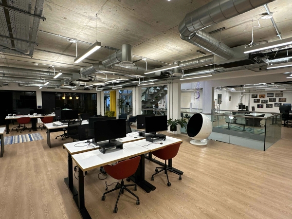 Bright open office space with industrial-style exposed ceiling and metal staircase, featuring desk spaces with orange chair and white egg chair, computer monitors