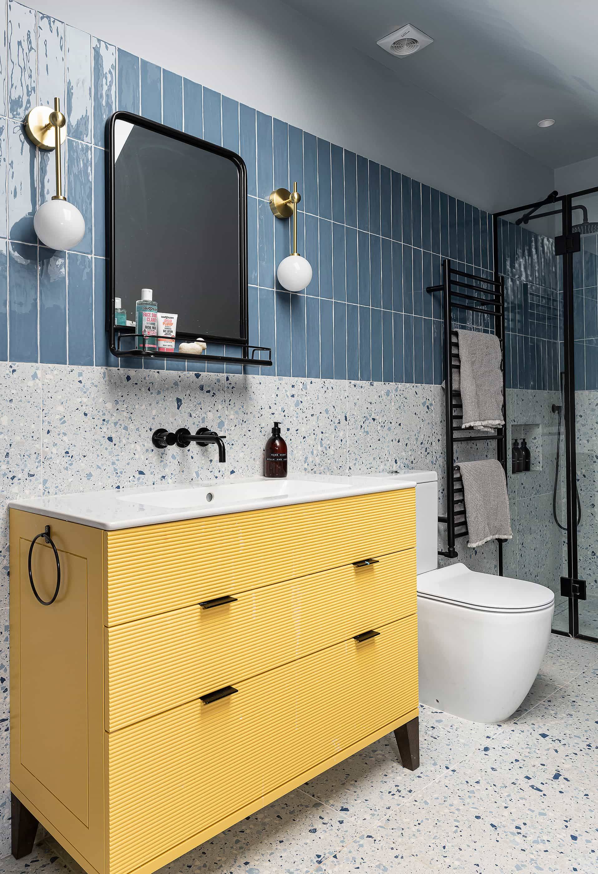 Modern bathroom featuring a yellow ribbed vanity cabinet, blue vertical tile splashback, and terrazzo wall tiles, ideal for skincare product marketing, Love Locations NW6.