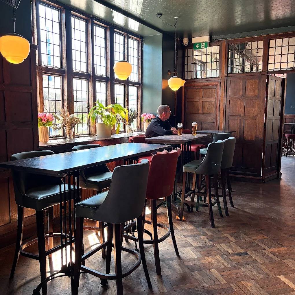 Cozy pub seating nook shoot space with high leather stools, dark timber panelling, and large leaded windows in Lewisham SE6 via Love Locations.