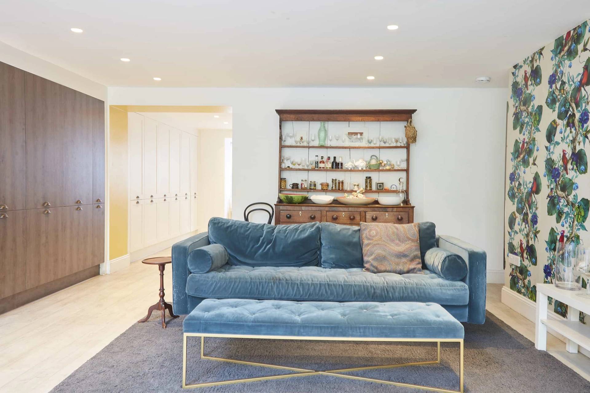 Symmetrical view of a blue velvet sofa and footstool set against white walls and antique wooden dresser shelving, Love Locations N5.