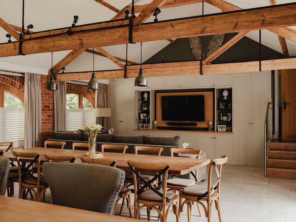 Open-plan living space in a converted Oxfordshire barn showing a large dining table, plush seating, and exposed timber framing, Love Locations.