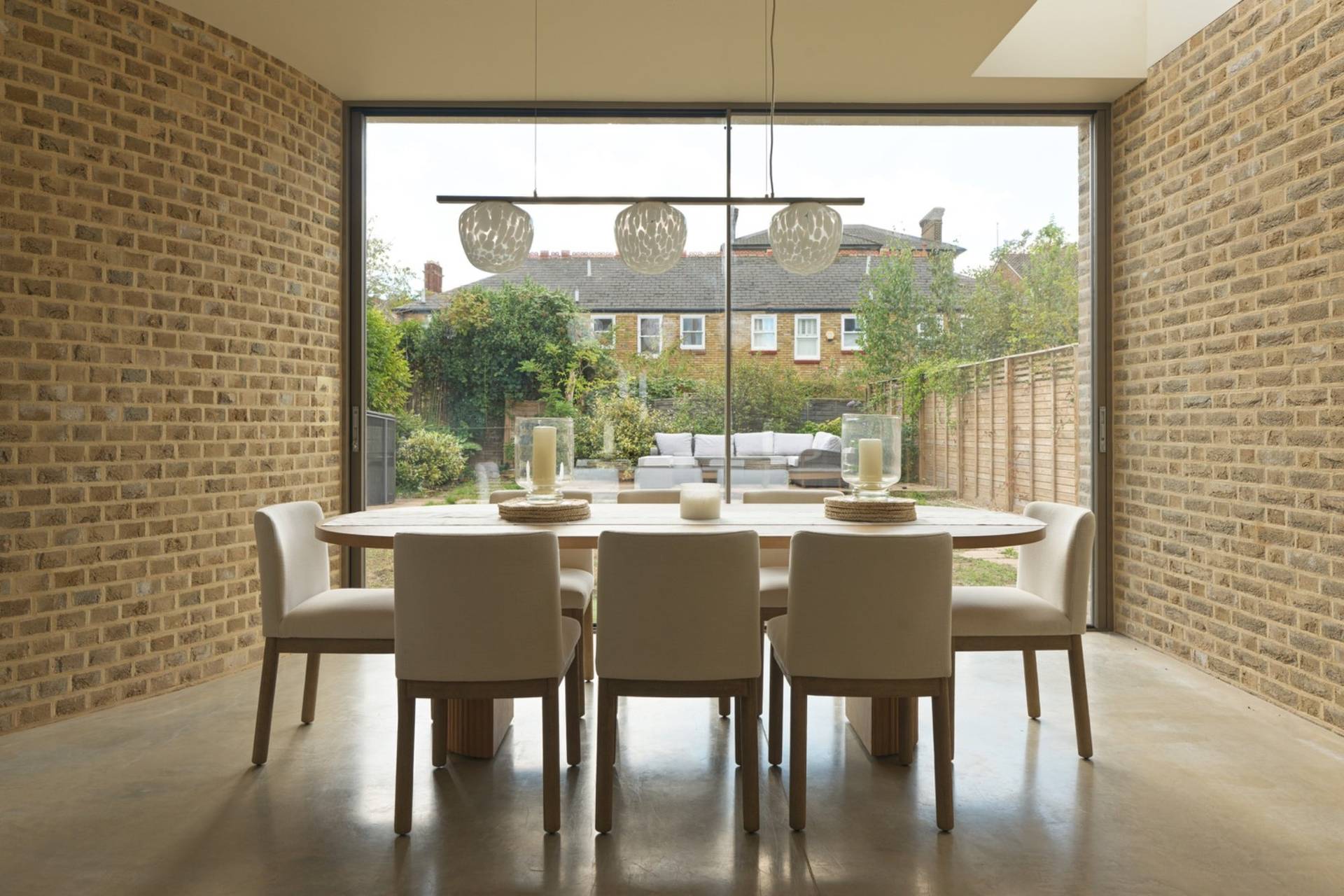 Elegant dining room shoot location featuring an oval wooden dining table, neutral chairs, exposed brick feature wall, and full-height glass doors by Love Locations.