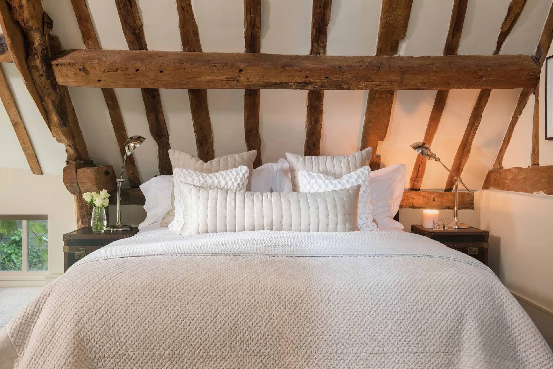 Close-up view of a cozy vaulted ceiling bedroom with a textured white quilted bedspread and dark oak ceiling beams by Love Locations.