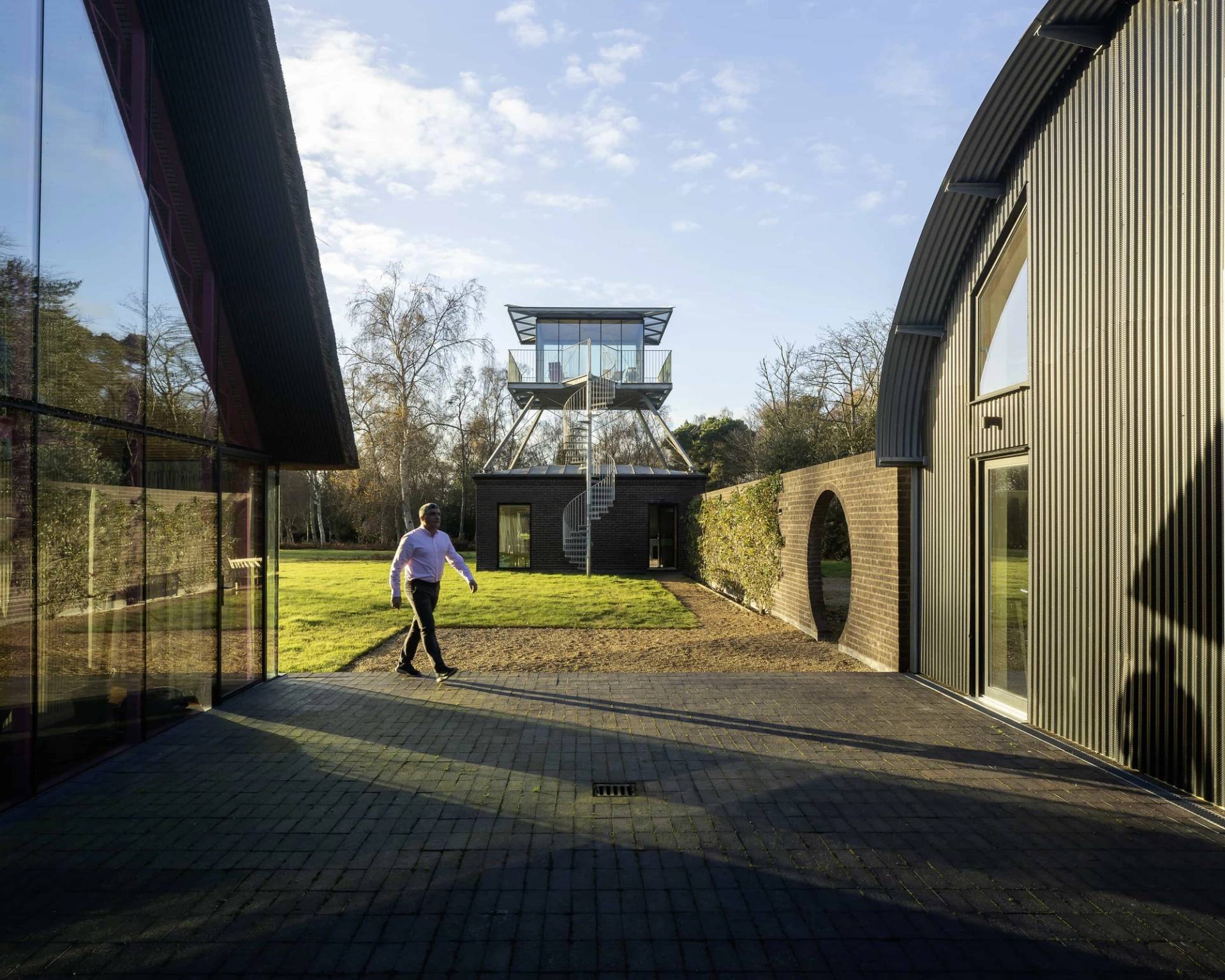 Courtyard pathway between modern black timber buildings leading towards an elevated glass tower, Love Locations IP17.