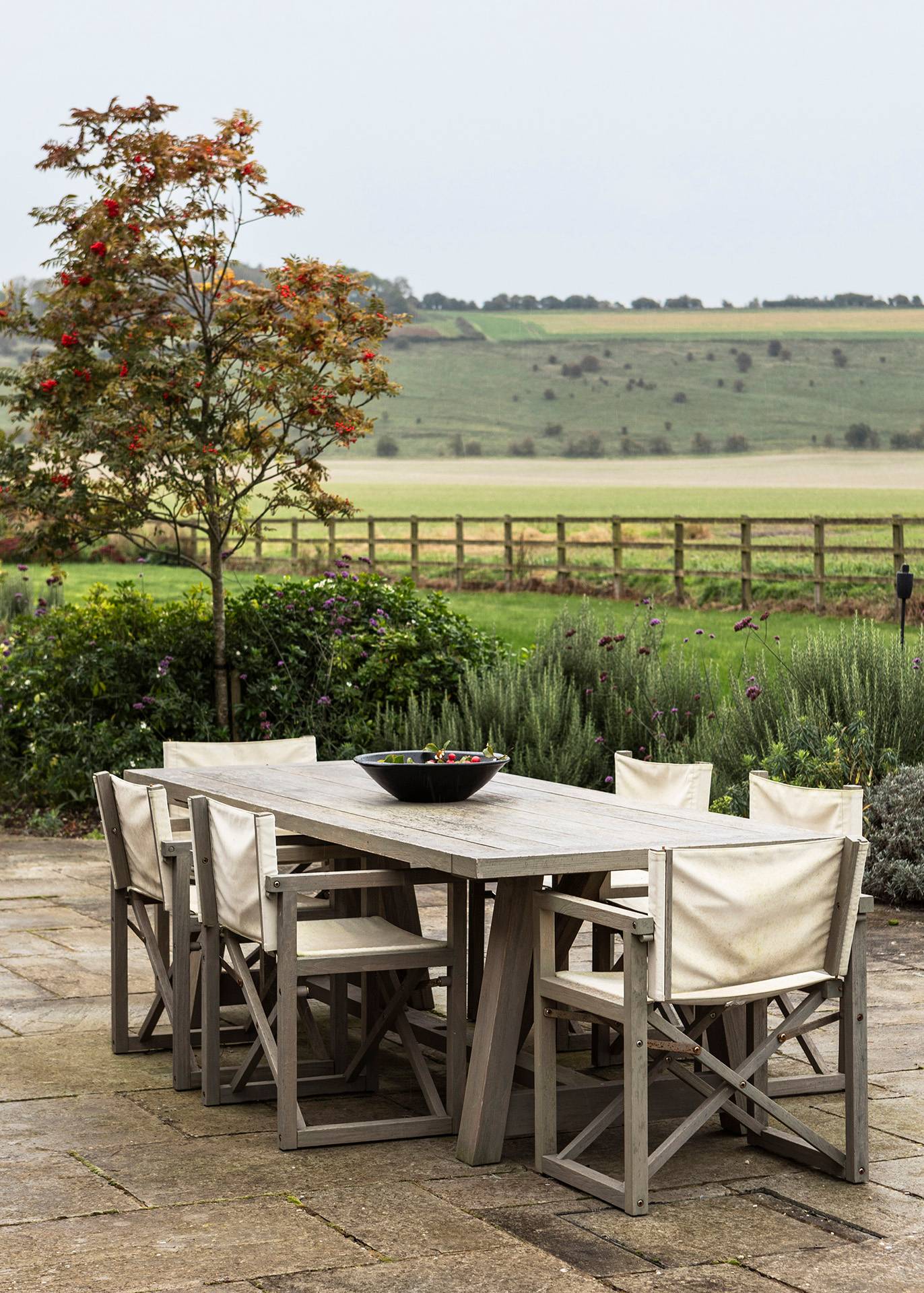 Outdoor teak dining table set with canvas chairs on a paved patio terrace surrounded by lavender, Love Locations SN8.