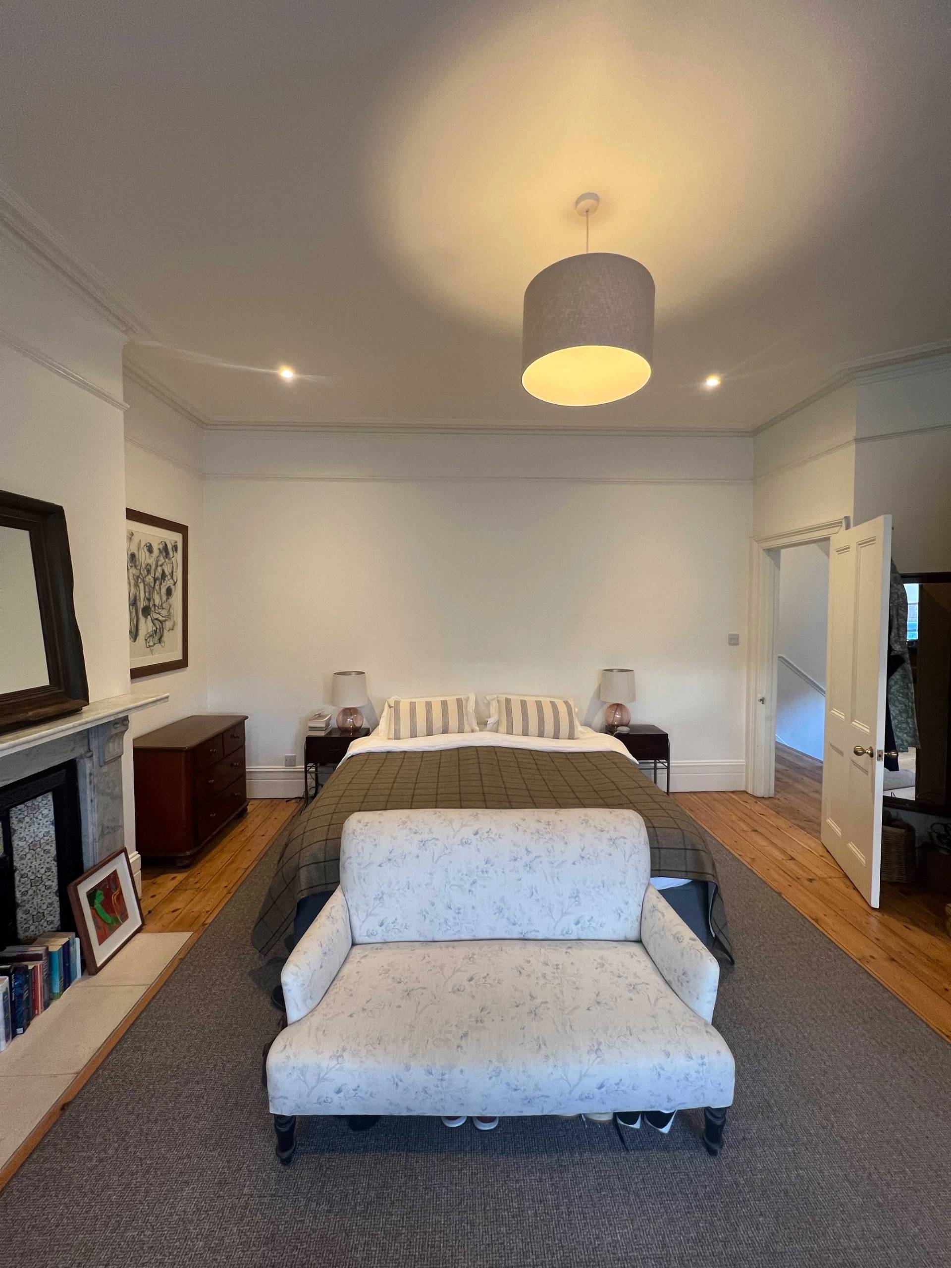 Symmetrical view of a master bed set against white walls with a grey bench and dark wood fireplace surround, Love Locations SE5.