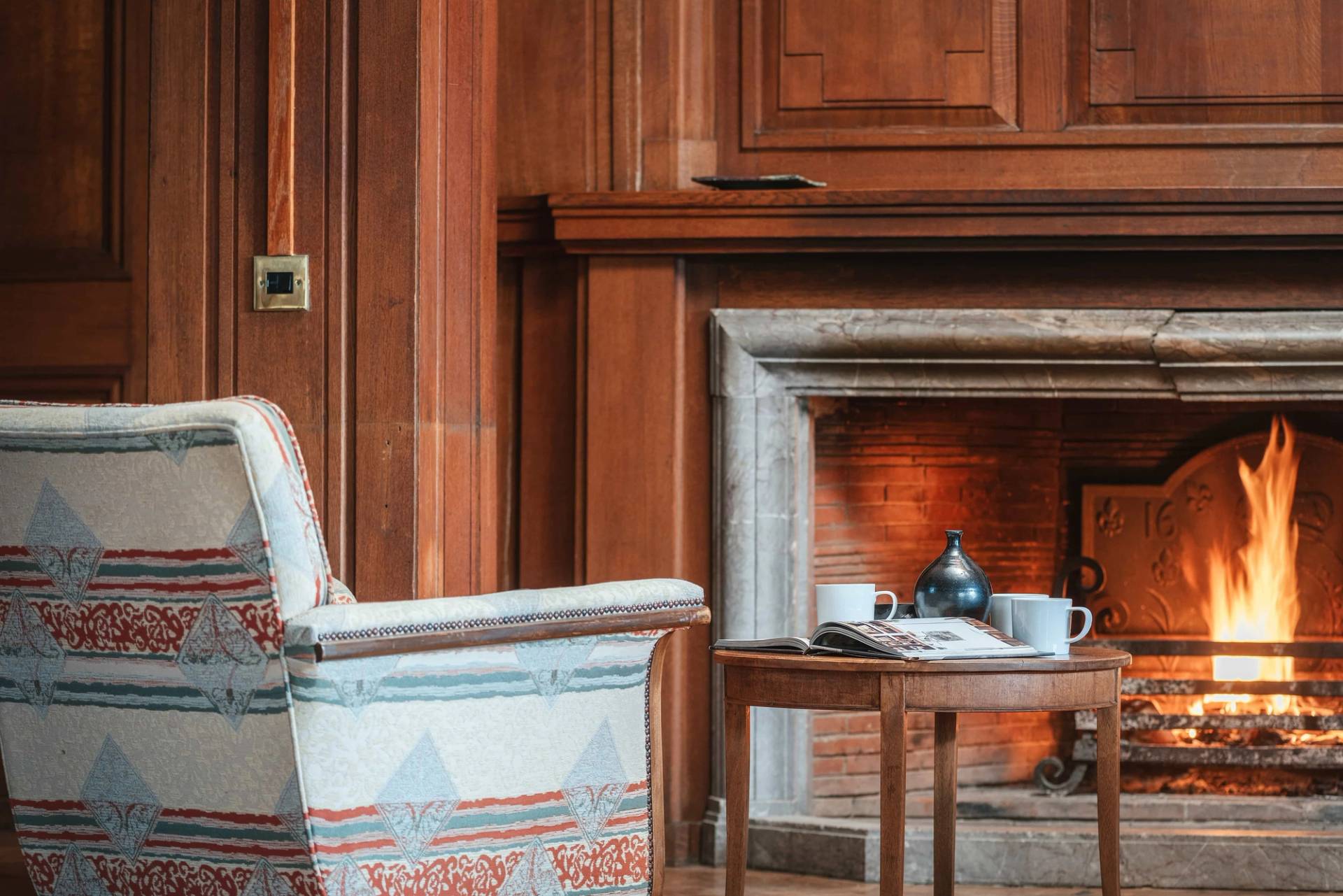 Close-up detail of a classic stone fireplace and armchair under wood panelling in a luxury country house through Love Locations.