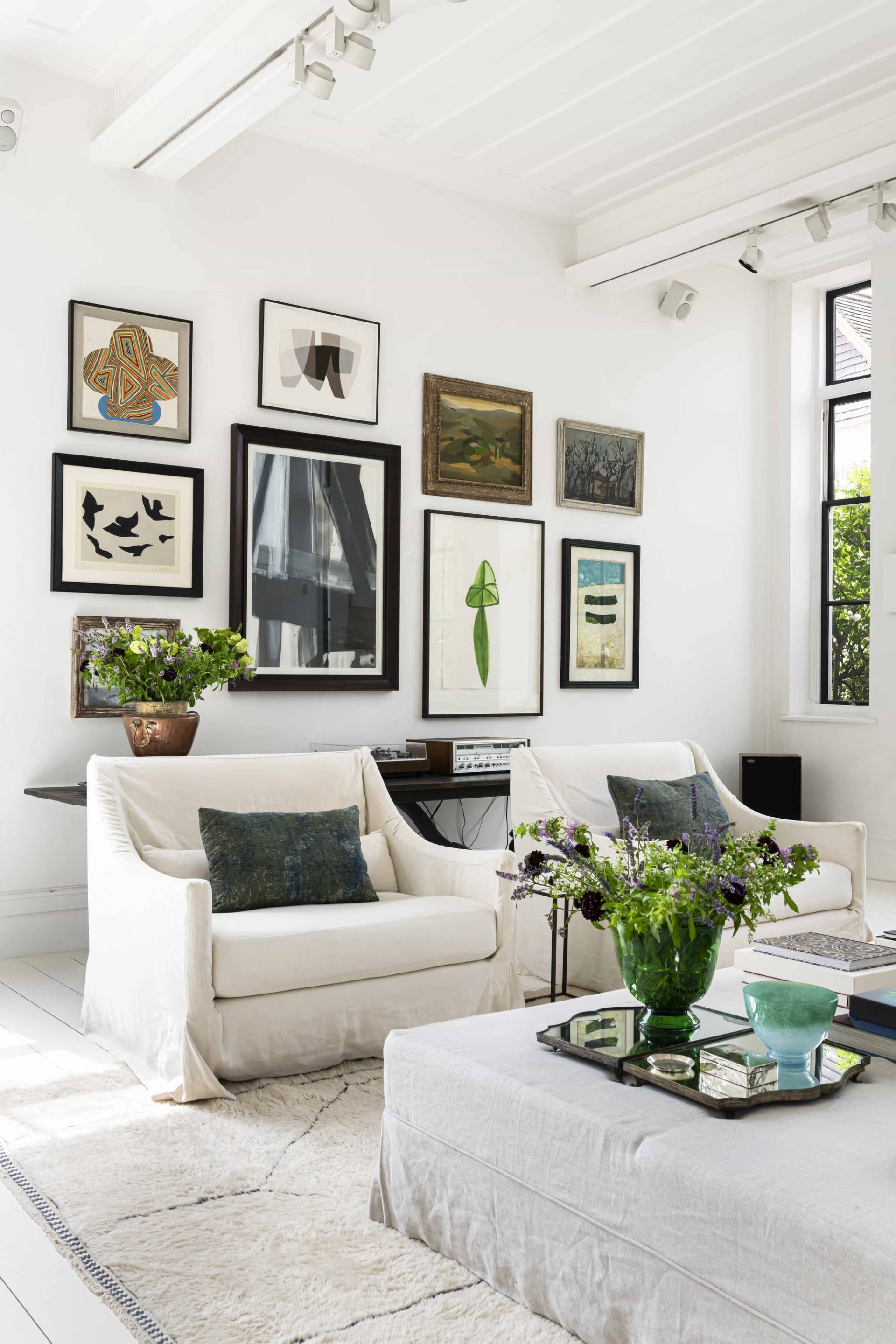 Cream armchairs and white sofa arrangement in a bright industrial studio apartment with tall Crittall windows and gallery art framing, Love Locations SW11.