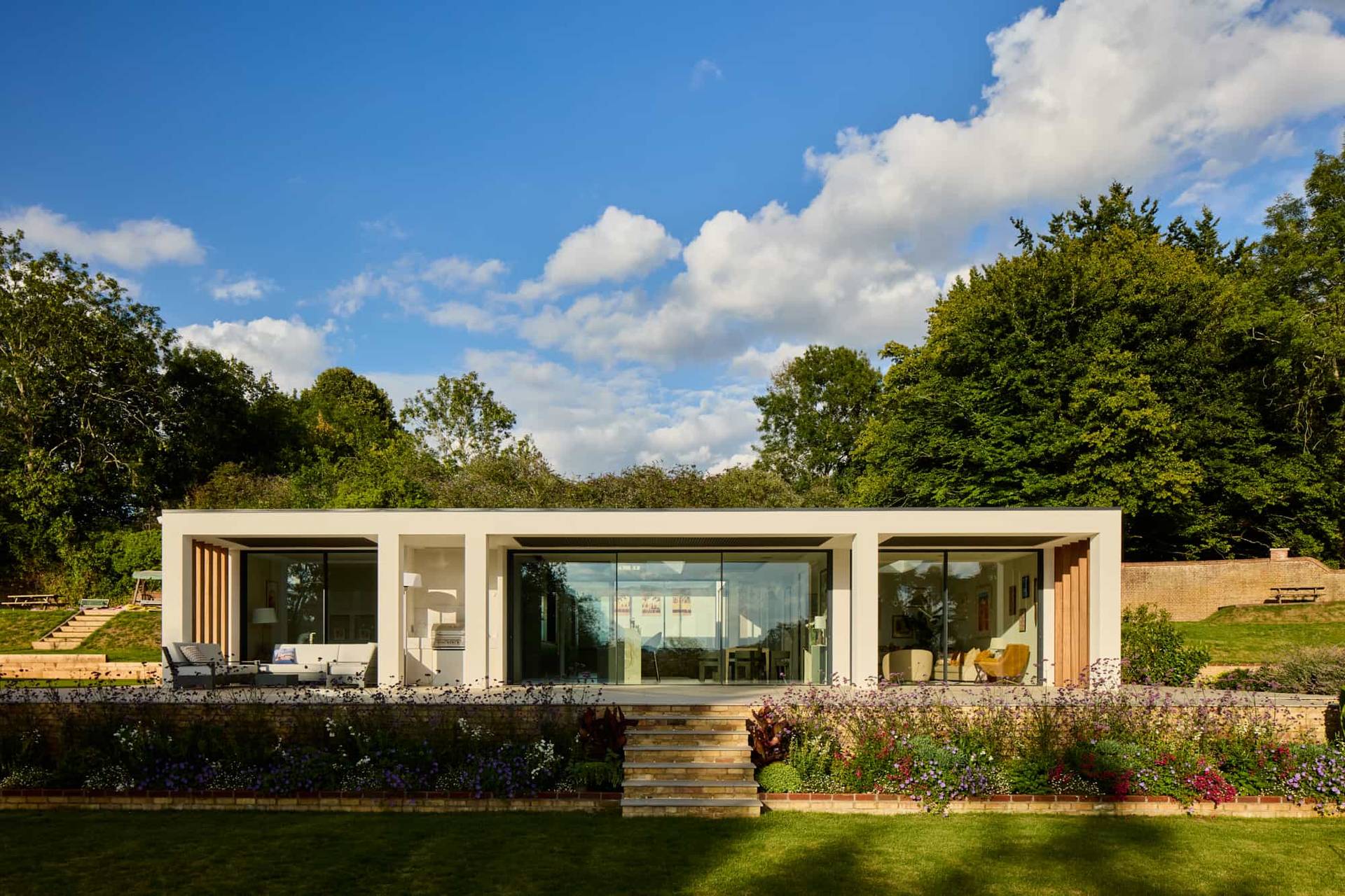 Rear architectural elevation of a modern white villa featuring glass sliding doors and timber accents set in lush grounds, Love Locations.