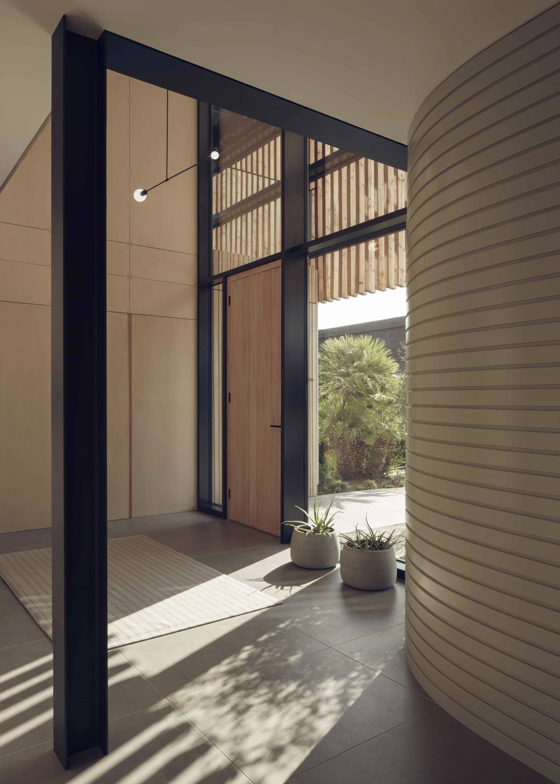 Architectural entrance hall featuring a solid pivot front door, light sandstone floor tiles, and a curved fluted wall, Love Locations NR9.