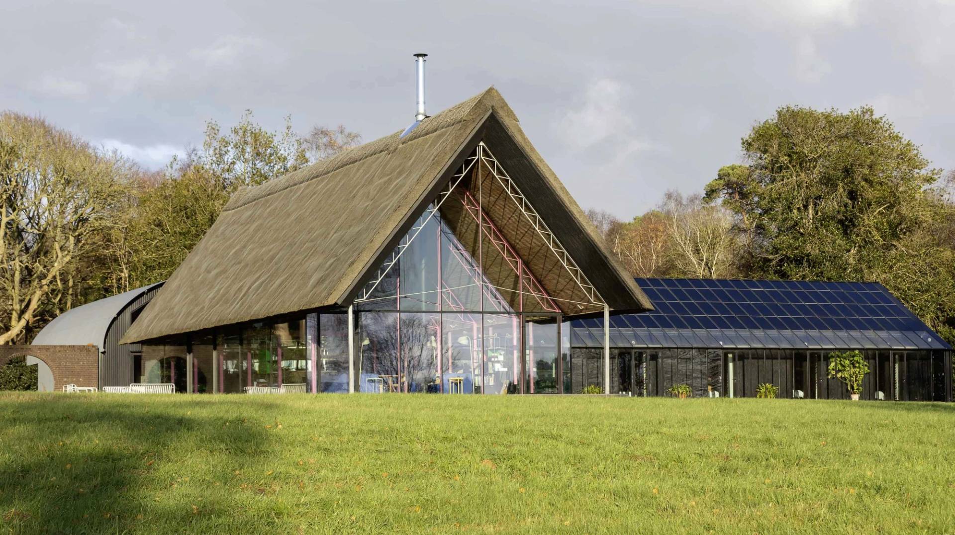Gabled front elevation of a modern thatched longhouse with full-height glass facade set on green lawns, Love Locations IP17.