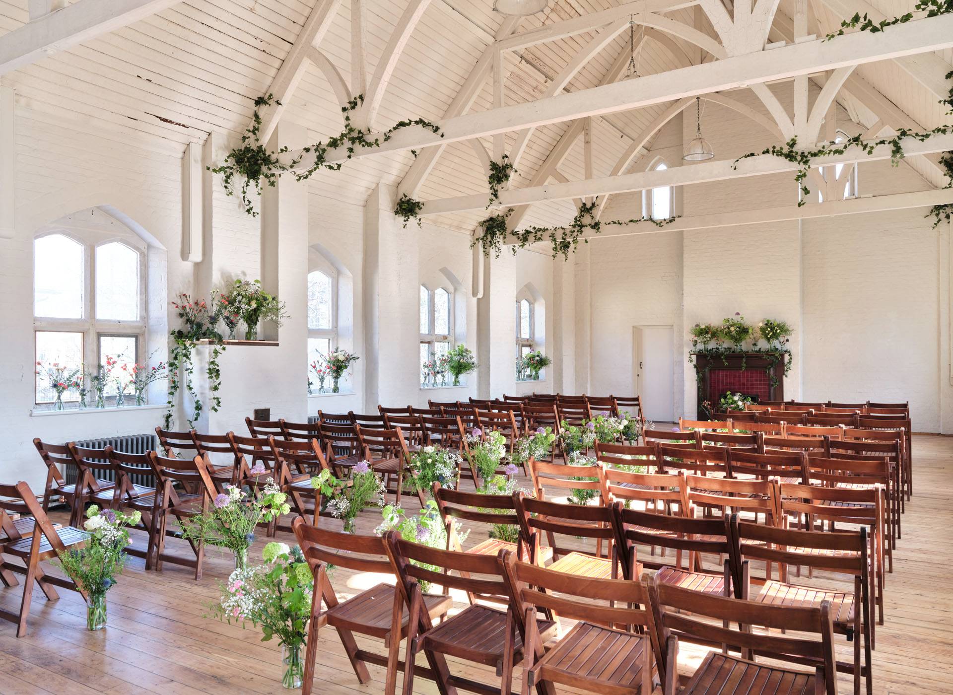 Wedding ceremony layout inside a white vaulted hall venue with wooden folding chairs and floral decorations, ideal for bridal campaign shoots, Love Locations E2.