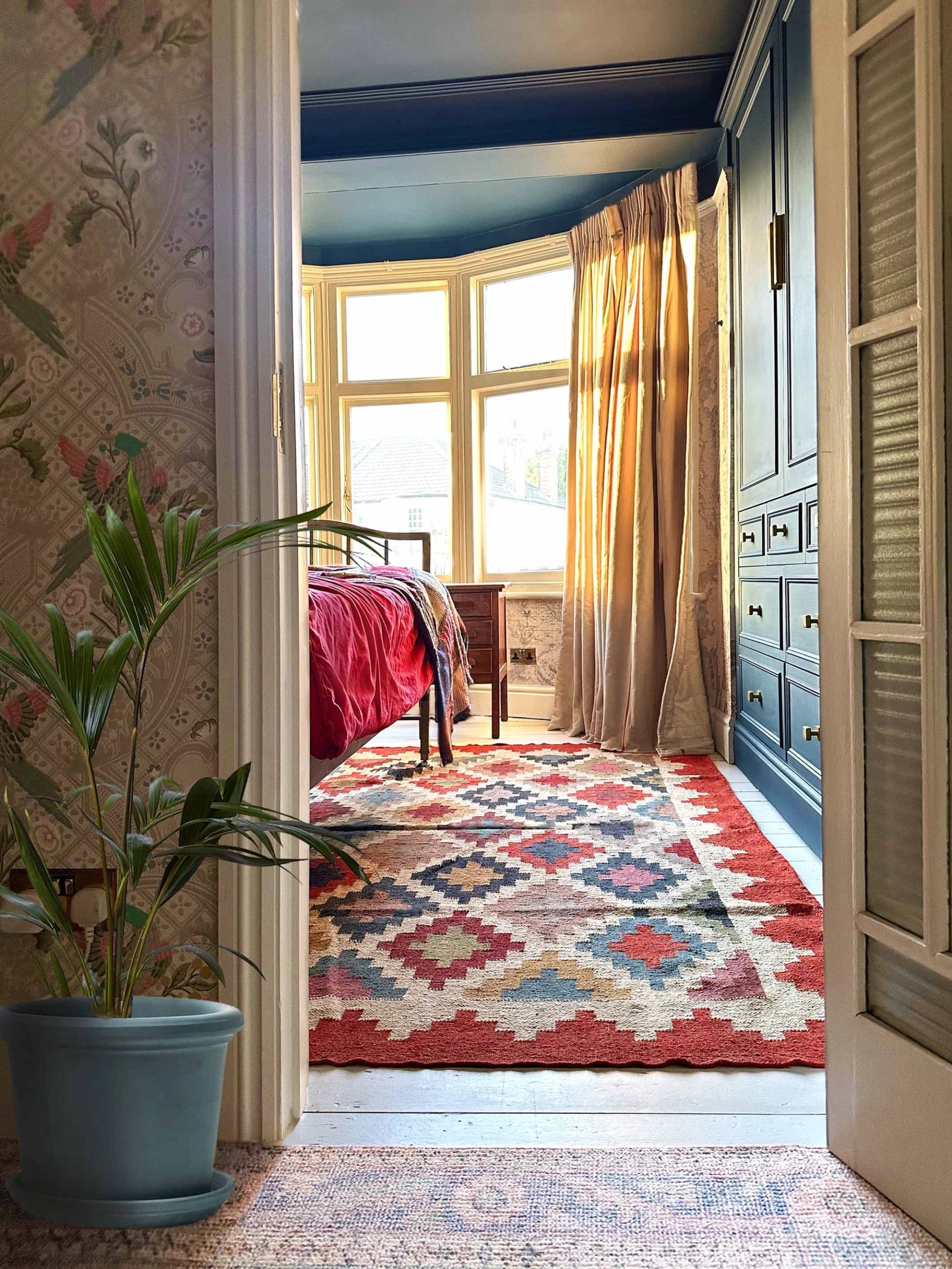 Bedside view through a timber door into a colorful bedroom with red rug and brass bed, Love Locations N13.
