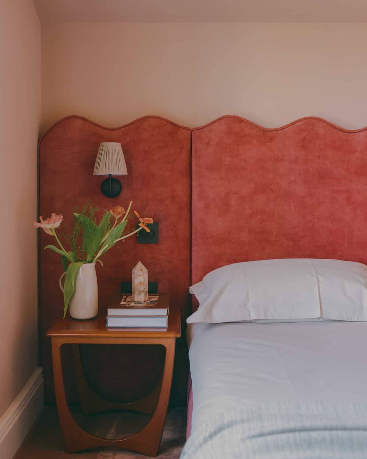 Bedroom detail featuring a scalloped terracotta red velvet headboard, wooden nightstand, and pink painted walls, Love Locations SW4.