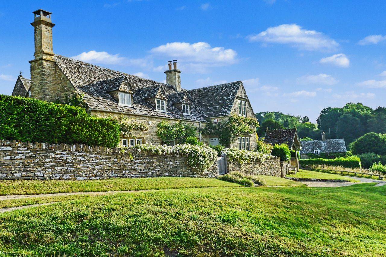 Period Cotswold stone cottage and church grounds film location in Gloucestershire GL7 via Love Locations, displaying manicured lawns, stone boundary walls, and classic English countryside architecture.