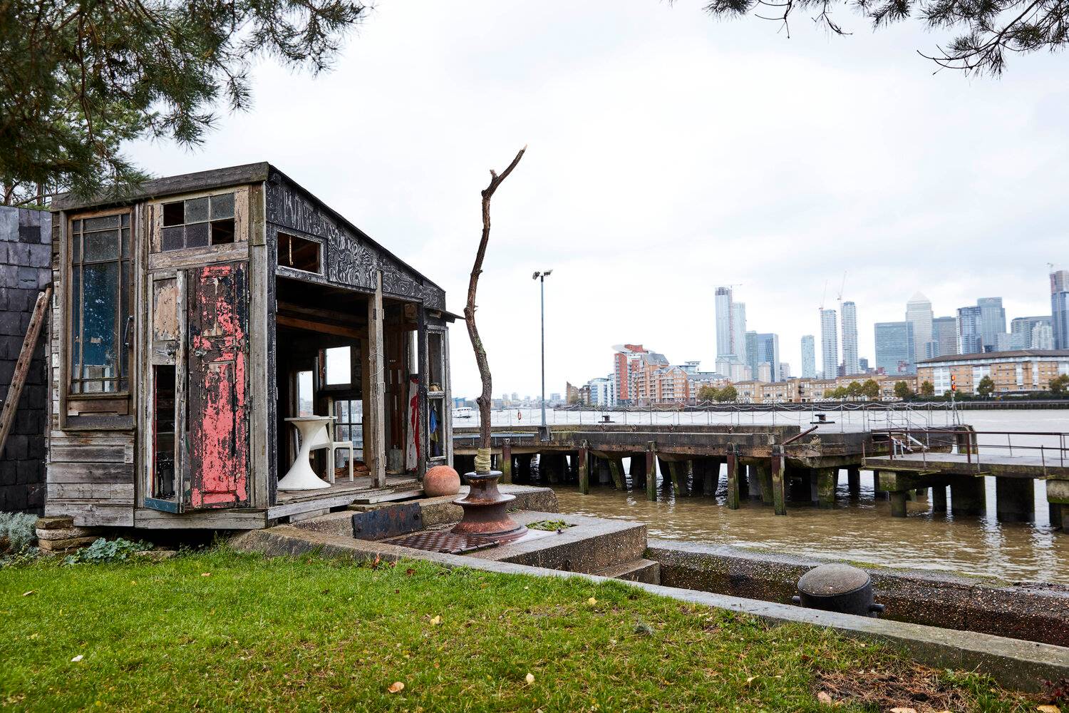 Riverside garden lawn looking out onto the River Thames and city skyline with a rustic wooden shack outbuilding, Love Locations SE8.