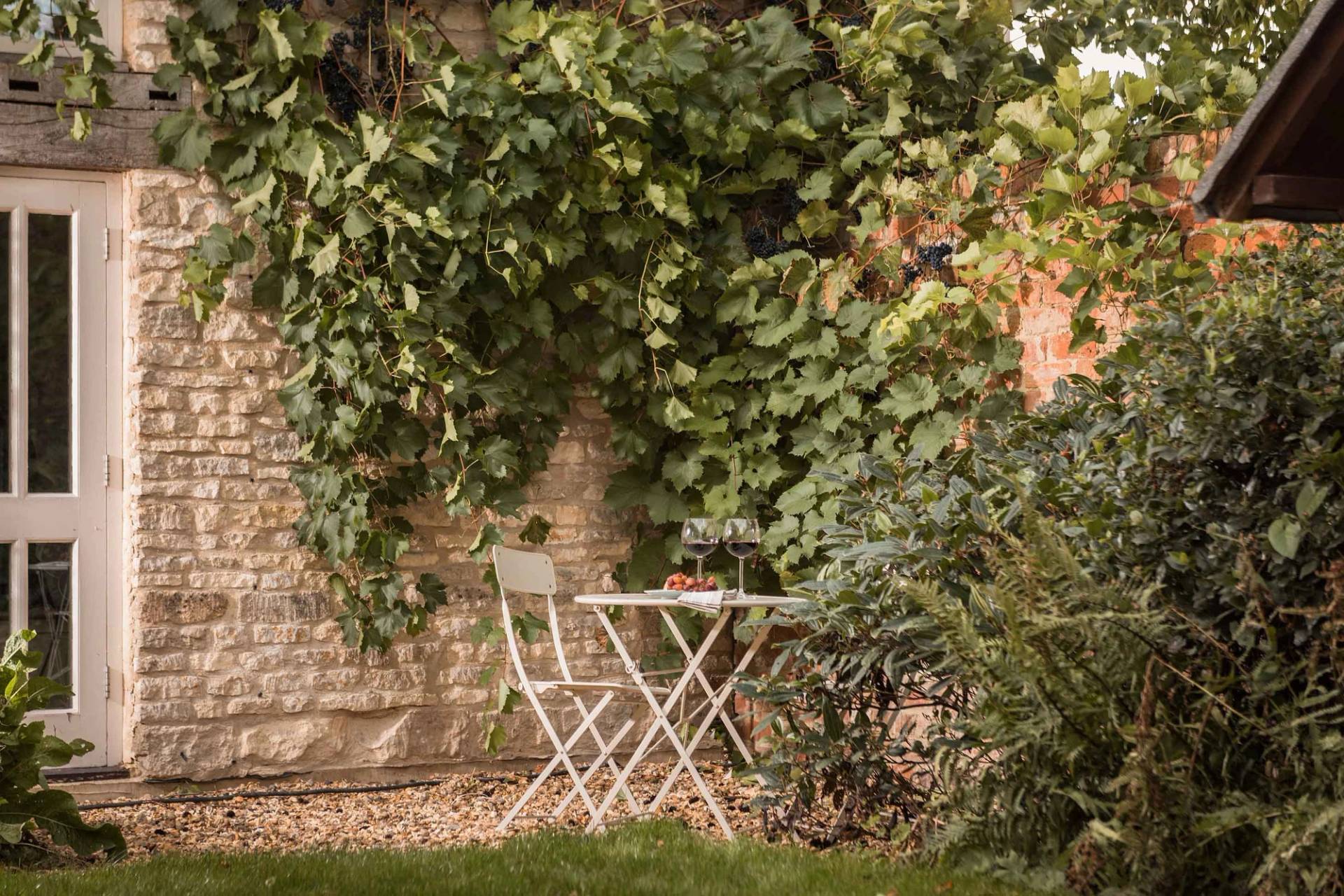 Intimate bistro seating nook set against a grapevine-covered rustic Cotswold stone wall by Love Locations.