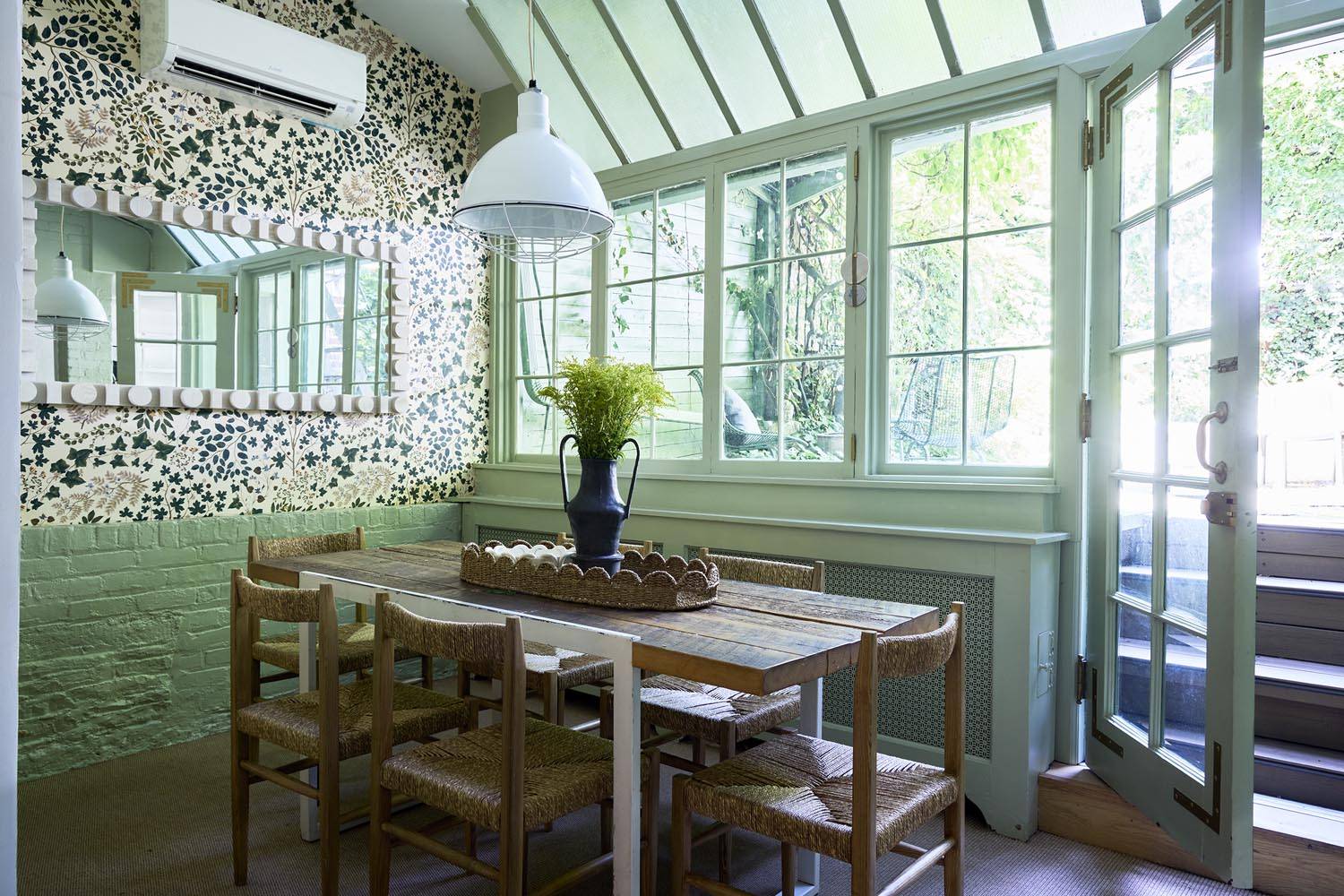 Sun-drenched garden room dining area in a Manhattan garden-level apartment from Love Locations, featuring a wooden table, woven chairs, and greenhouse glass windows looking onto lush foliage.