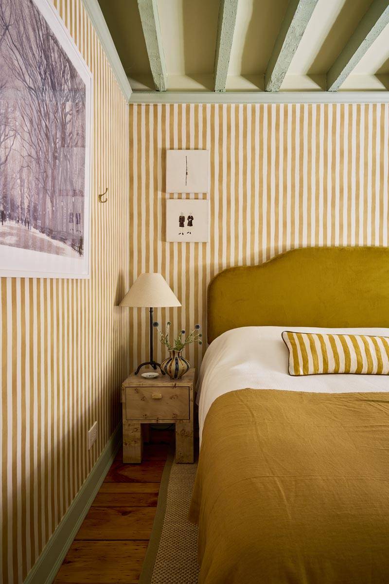 Detail shot of a mustard velvet upholstered bed and bedside table in a stripe-wallpapered bedroom shoot space in Manhattan via Love Locations.