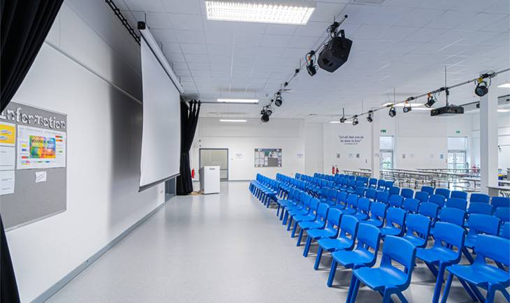 Spacious school event hall shoot space with white floors, overhead stage lights, retractable screen, and blue seating via Love Locations.